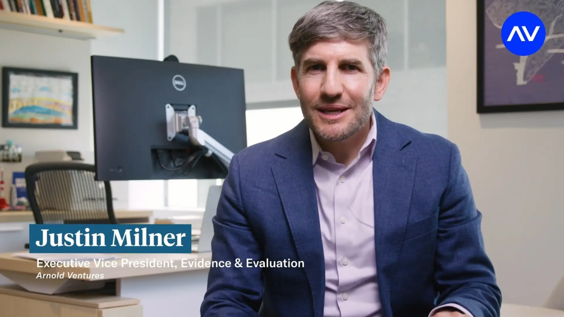 Person in a blue blazer speaking in an office, with a monitor behind them and a lower-third reading “Justin Milner, Executive Vice President.”