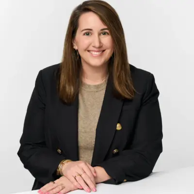 A smiling woman with long brown hair wears a black blazer over a beige top, resting her hands on a white surface.