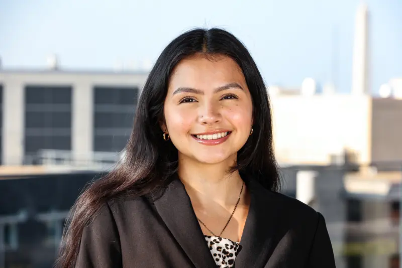 A young woman with long dark hair smiles warmly in a business suit, with a city skyline and blue sky in the background.