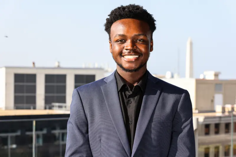 A young man with a bright smile wears a dark suit jacket over a black shirt, standing against a cityscape background.