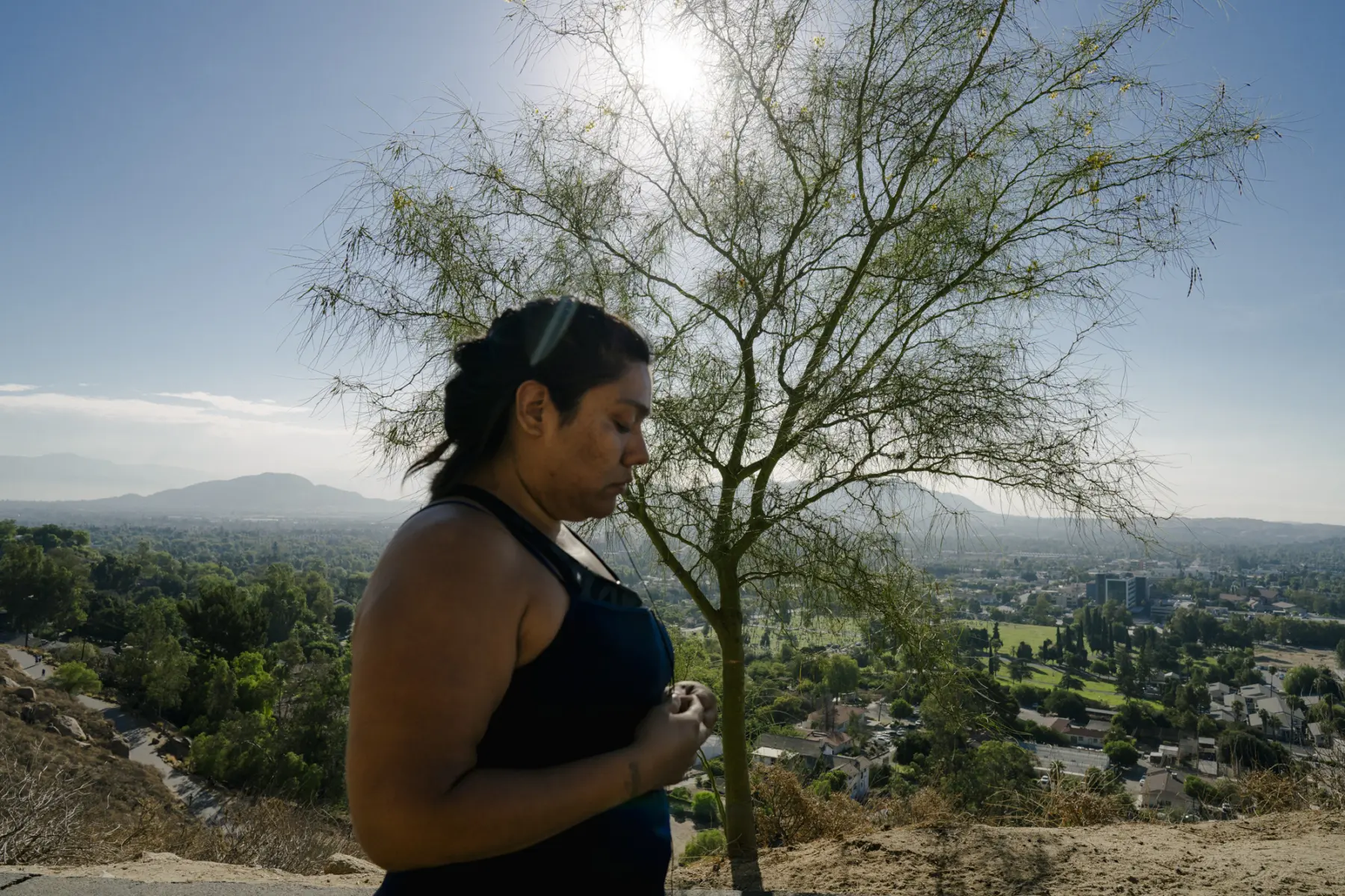 Melissa, a Hispanic woman, stands in front of a tree on a hill overlooking a city landscape. She is out of focus. The tree and landscape are in focus.