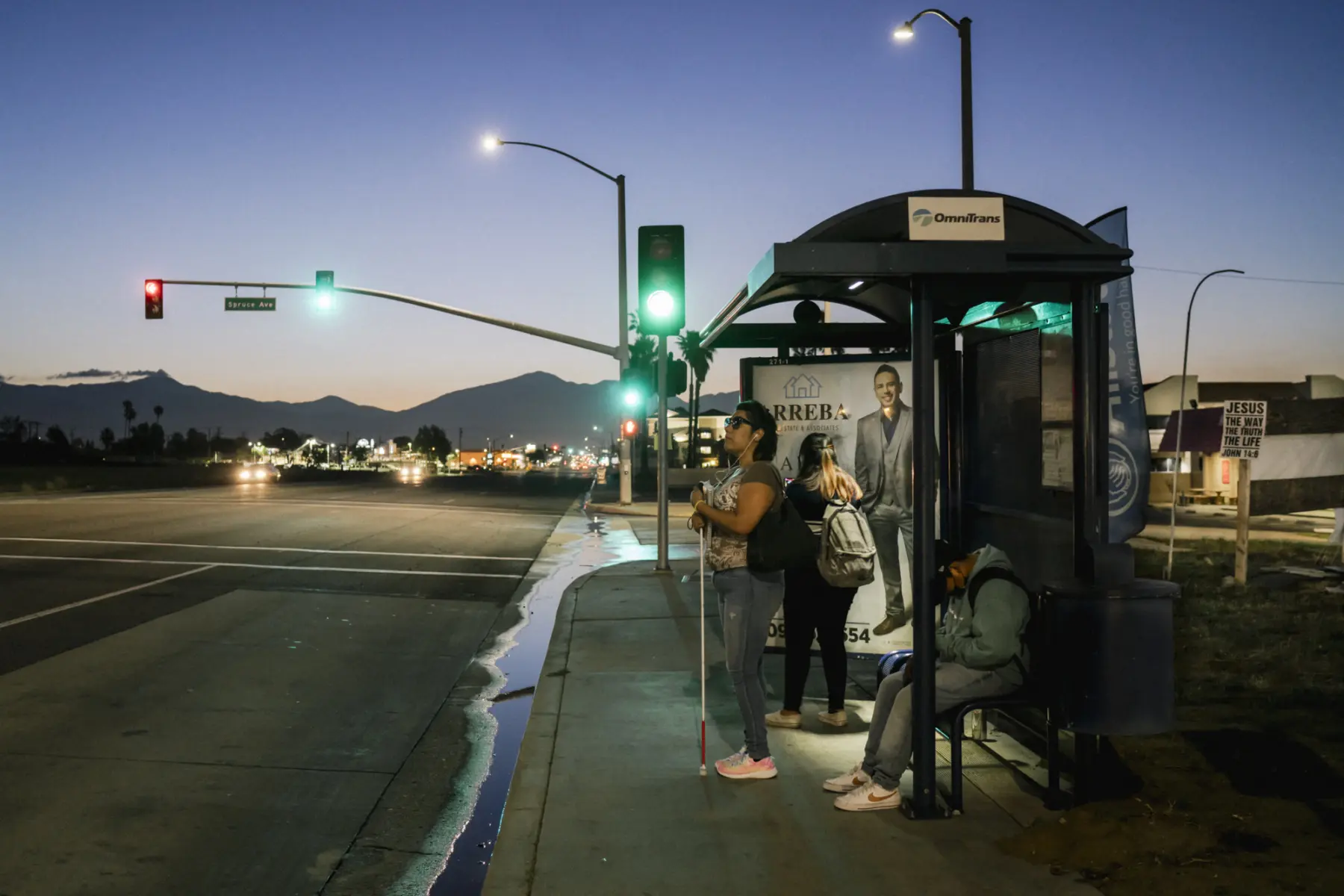 Melissa stands, holding her white cane, at a bus stop shelter next to an empty street and traffic light. Her teenage children, Angie and Jacob, stand and sit beside her wearing backpacks. The sun rises over the outline of a mountain range behind them.