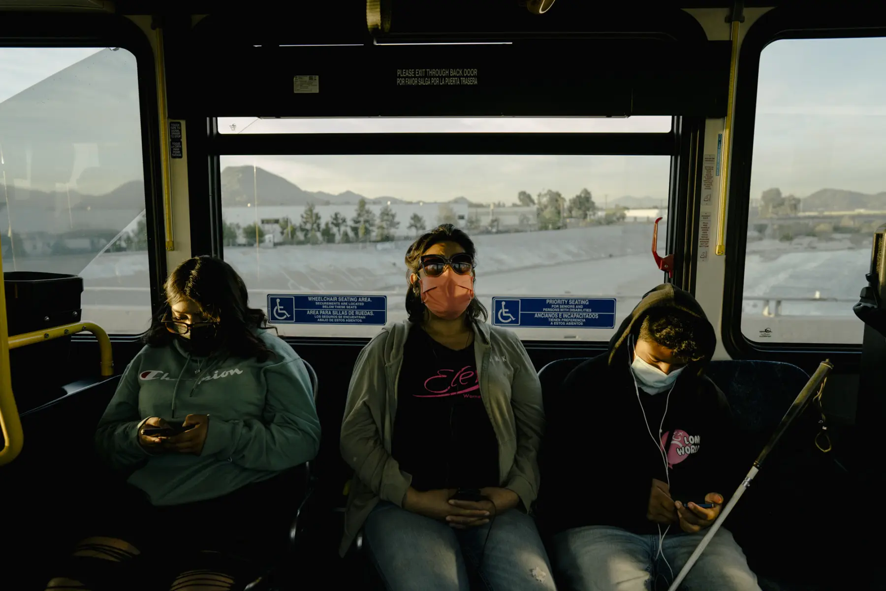 Melissa wears sunglasses and listens to music on earbuds between Angie and Jacob, who look at their phones.The landscape and mountain range are seen through a window behind them. Light hits their faces.