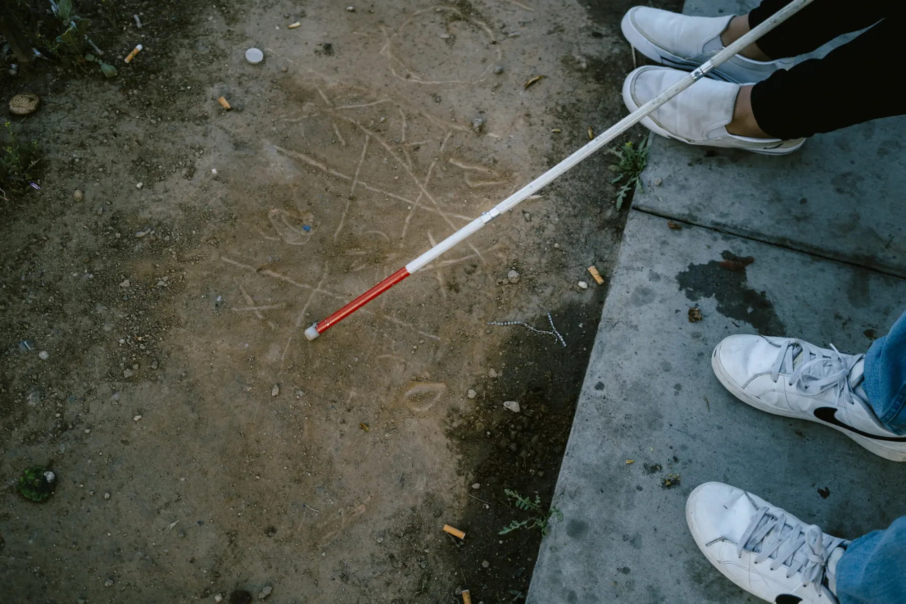 Melissa’s white cane rests on a game of tic-tac-toe etched into dirt next to a concrete sidewalk, where Angie’s and Jacob’s sneakers are seen side-by-side.