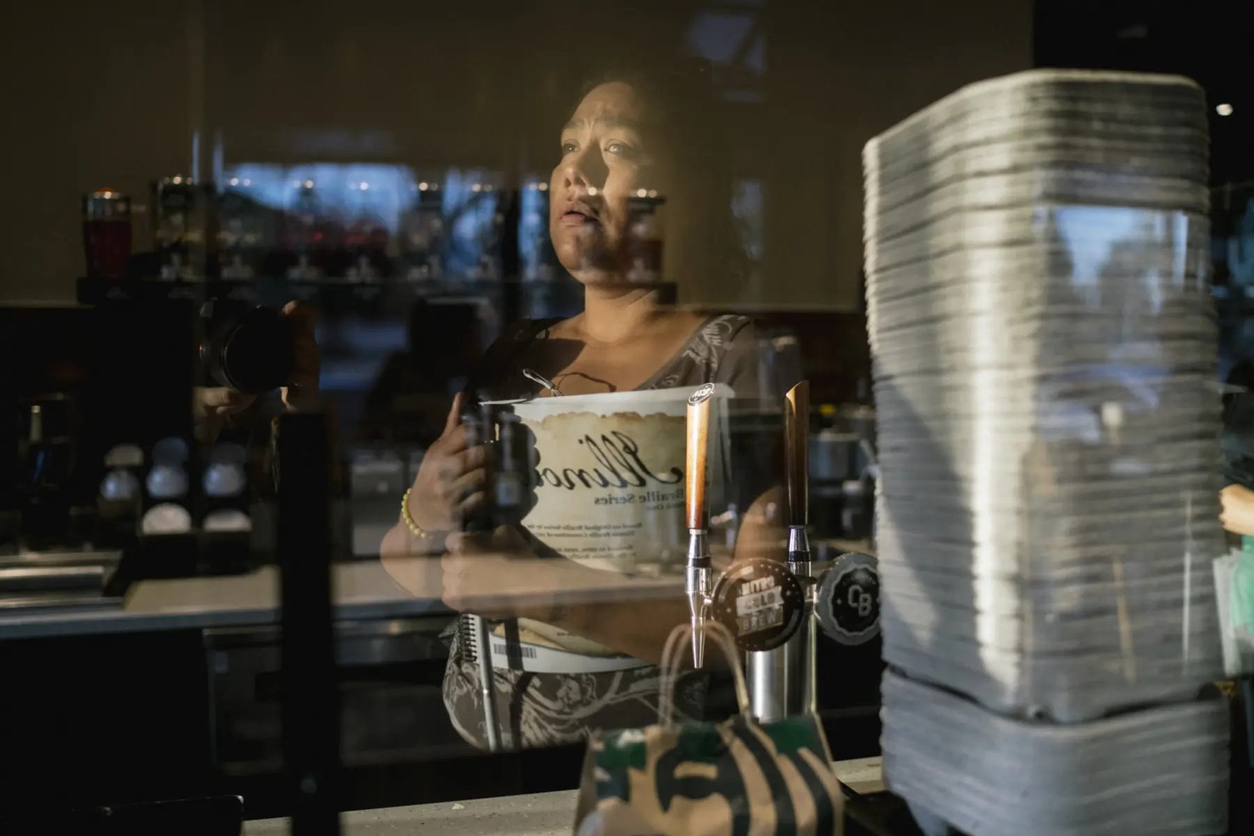 Melissa is reflected in a glass divider at Starbucks coffee. She holds her Braille book to her chest as morning light shines on her face and torso.