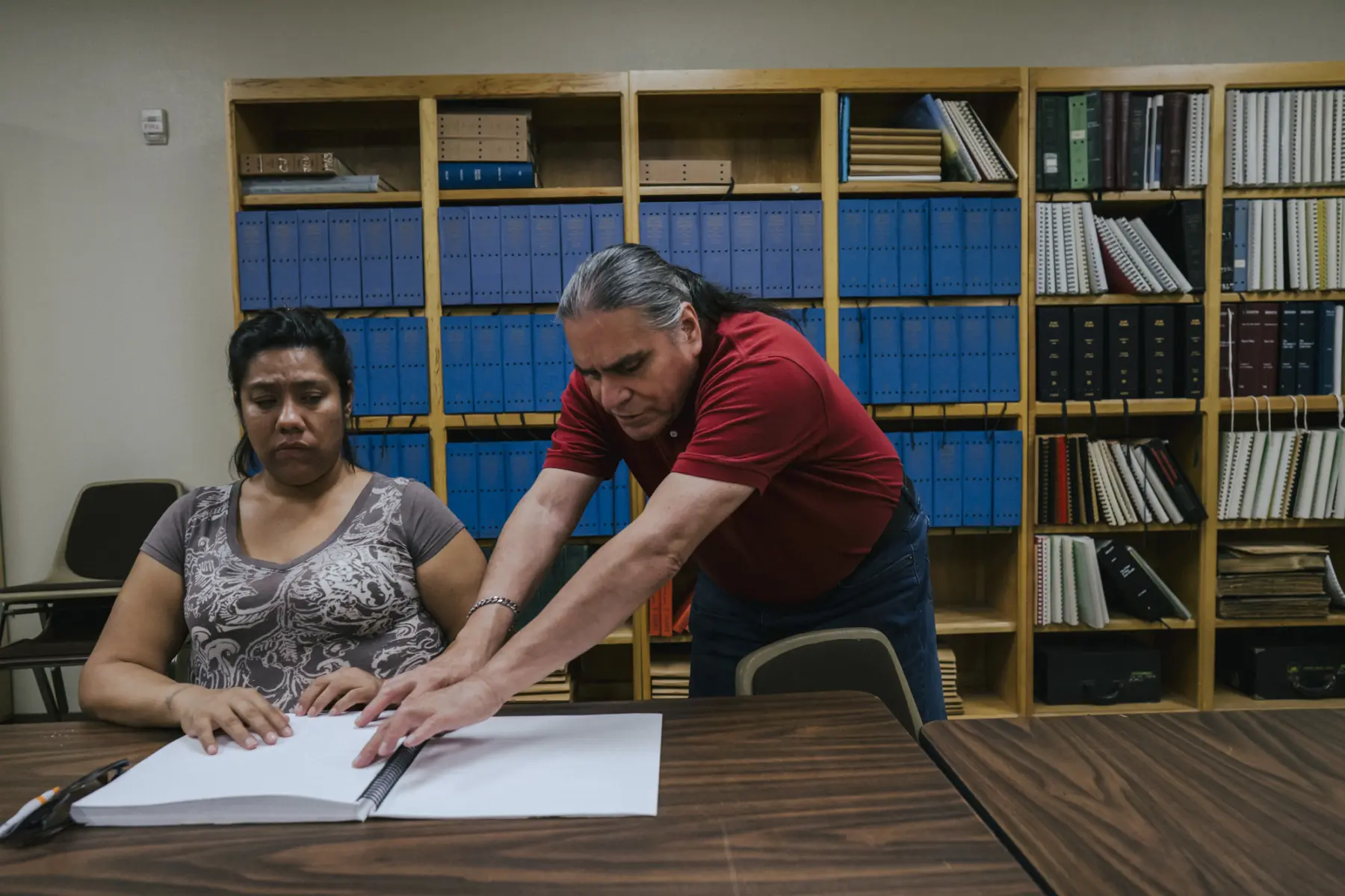 Melissa sits at a conference table with hands on her Braille book. Arturo, an older man with a gray ponytail who is also blind, reads Braille from the same page as he stands next to Melissa. A large case of books is behind them.