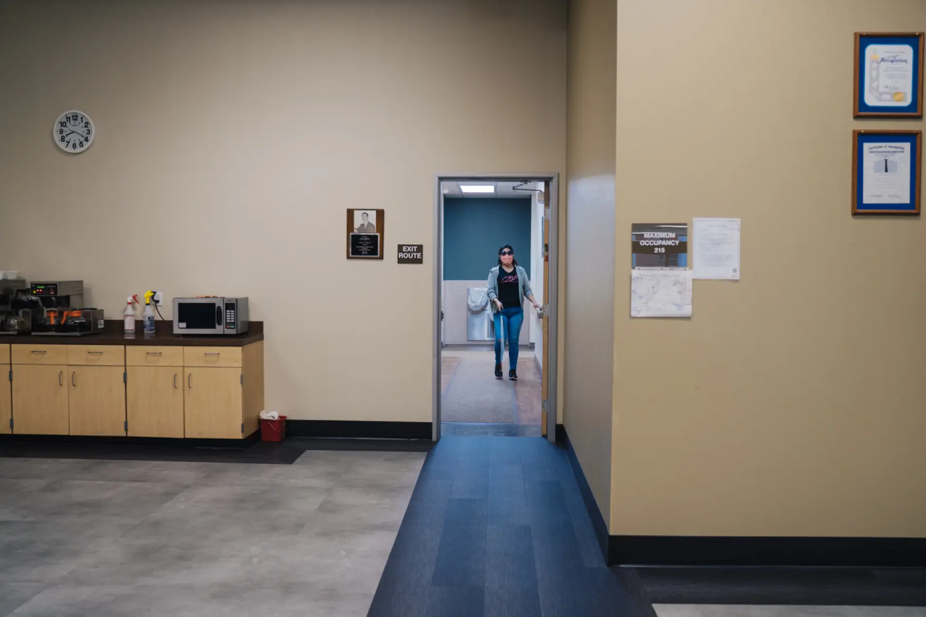 Melissa is seen in the distance as she holds onto a wall while making her way toward a doorway inside an office-like building.
