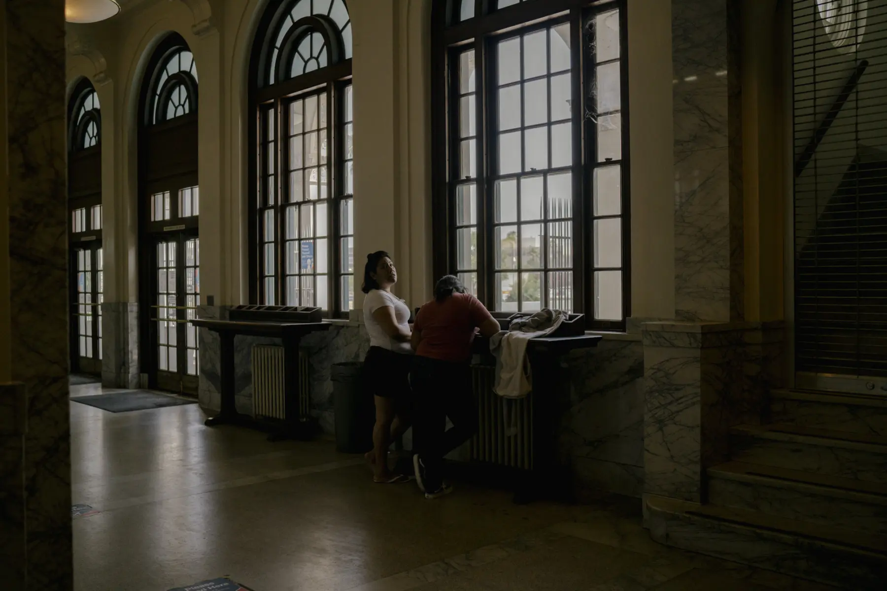 Melissa stands next to Angie in front of a counter at a dimly lit old post office with large glass windows. Melissa is turned toward Angie, who leans against the counter with her head down.