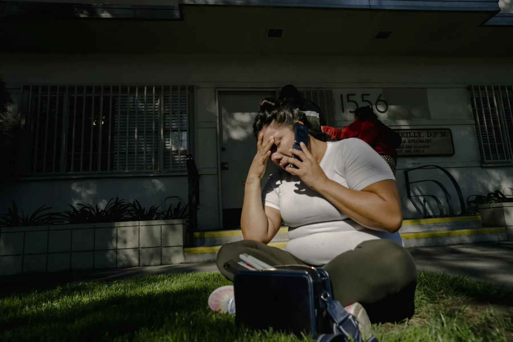 Melissa who sits cross legged on a patch of grass in front of a doctor’s office. She holds her phone to her ear and rests her head on her hand