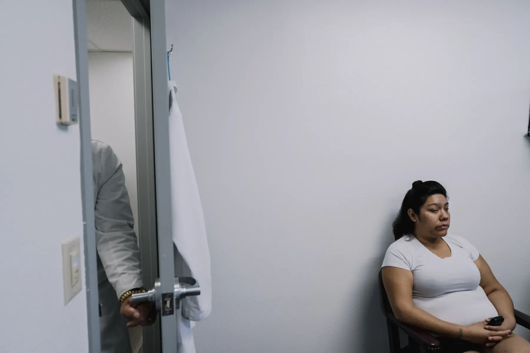 Melissa sits patiently in a doctor’s office against a stark white wall. A door opens to the left revealing only a doctor’s arm pushing the door open toward her.