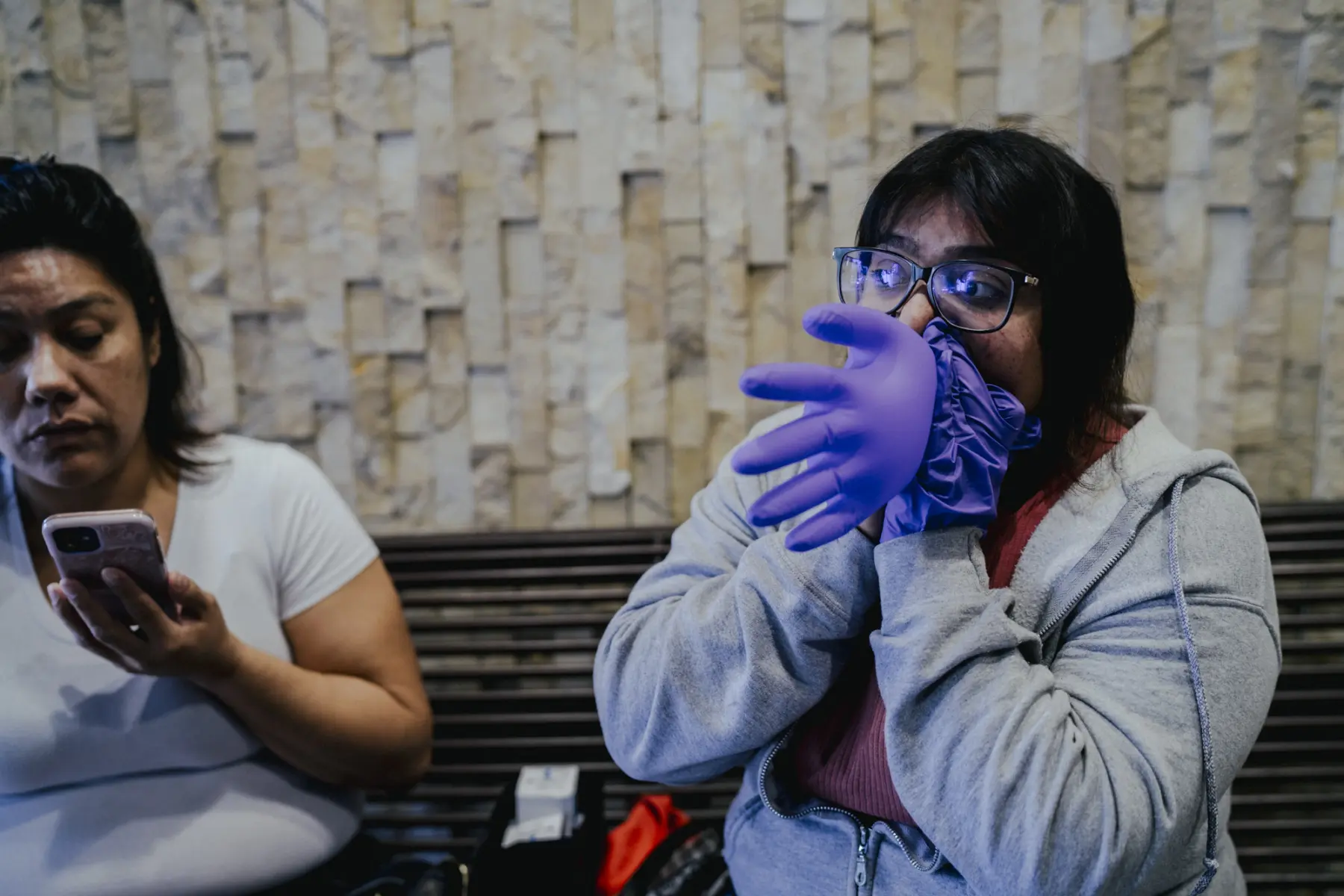 Angie looks to the side as she blows air into a purple medical glove, making it into a balloon. Melissa, out of focus, sits next to her holding a smartphone.