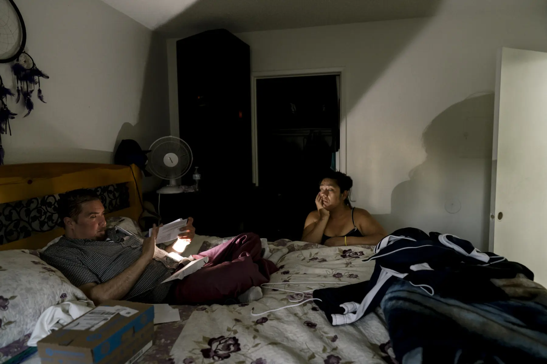 Melissa kneels next to a bed while Scott points a smartphone app with flashlight onto a document as he sits on the bed. The flashlight creates a large shadow of Melissa on the white wall behind her.