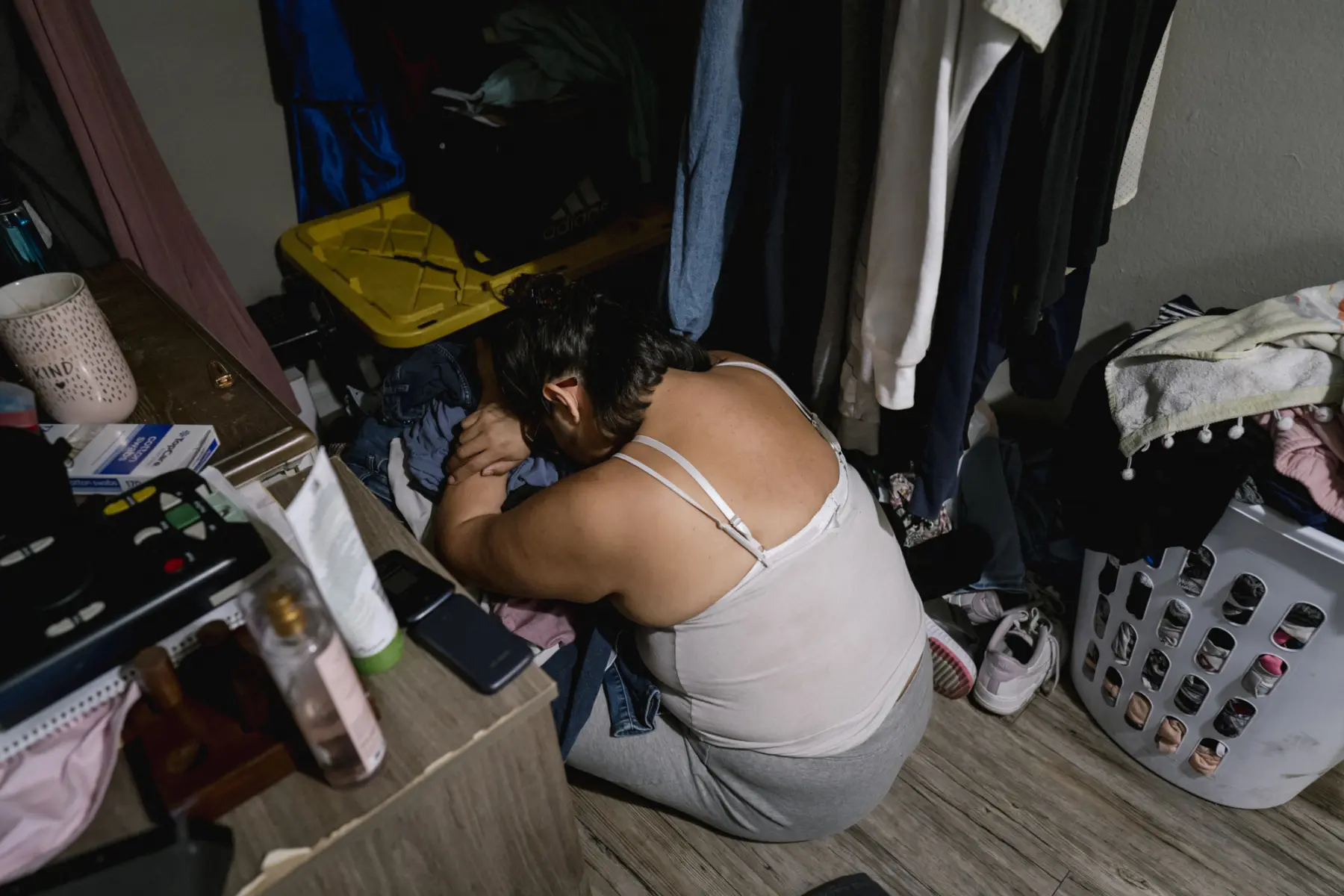 Melissa kneels on the floor resting her head on a pile of clothes in a closet. Clothes hang on one side of her body. On the other side, personal items like perfumes, a candle, and a reader are seen on a dresser.