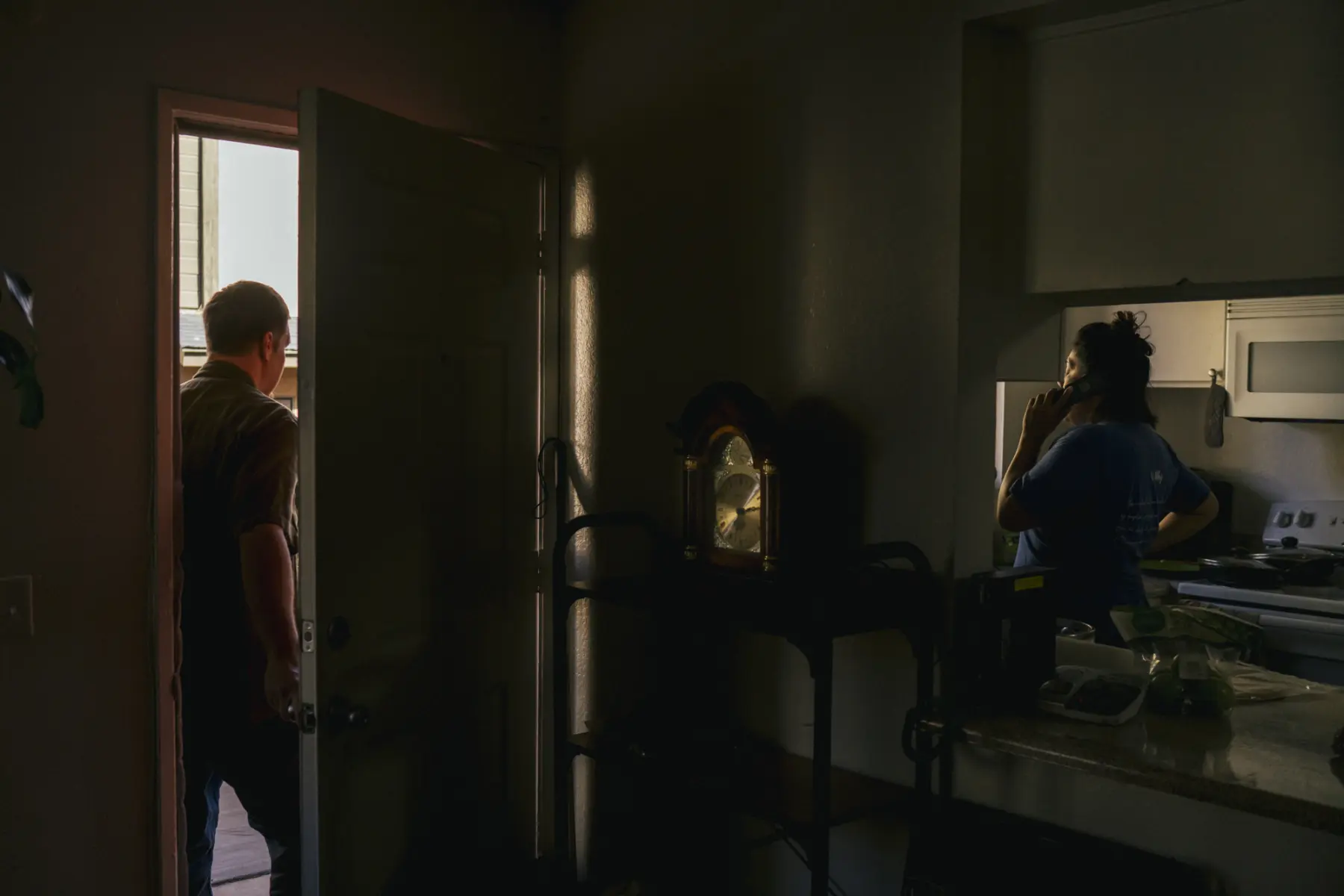 Melissa stands in the dimly lit kitchen on the phone. Scott stands in the front doorway. They are divided by a wall with a large classic clock displayed on a table in front of the wall.