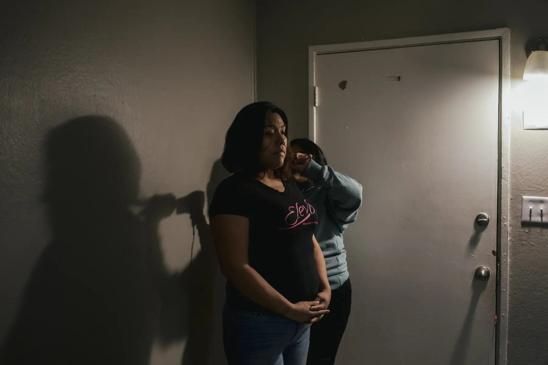 Melissa stands by a door while Angie stands behind her blow drying her hair. Shadows of Melissa’s figure and Angie’s raised arm are seen on the wall beside them.