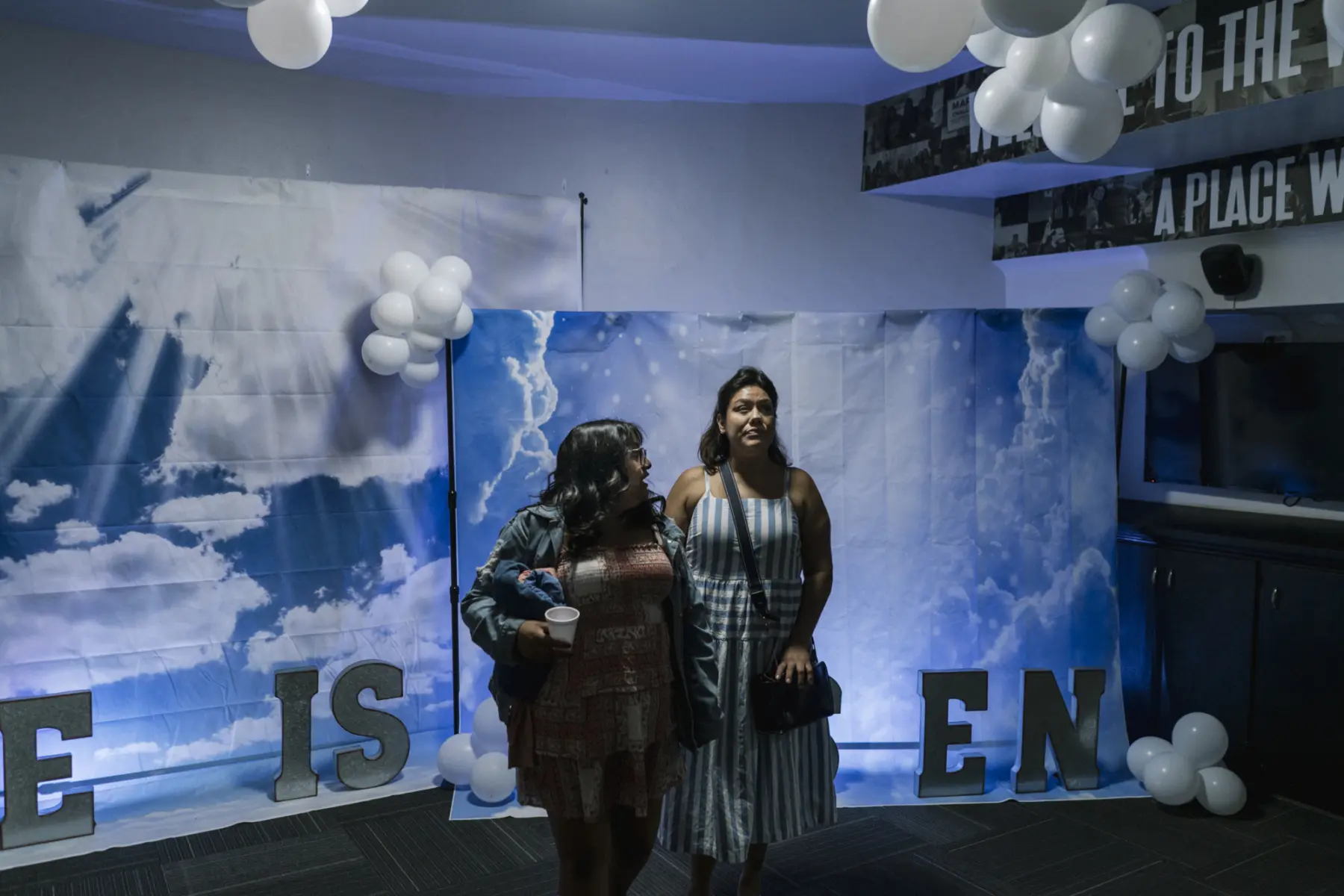 Angie looks to her mother, Melissa, as they walk arm in arm in front of backdrops of blue skies and large clouds. A portion of large letters below the backdrops read “He is risen.” White balloons hang from the ceiling and from the backdrops.