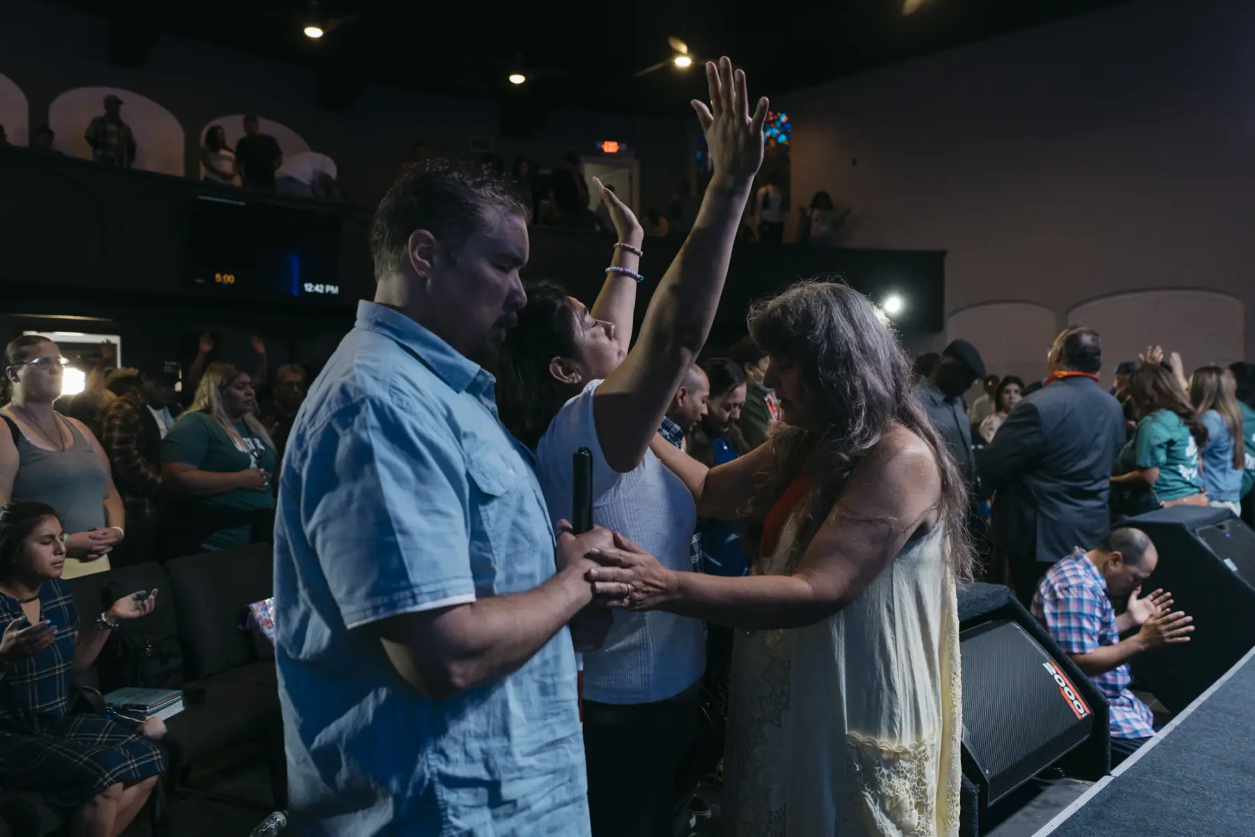 Melissa holds her hands in the air and closes her eyes as she receives a prayer at a church altar. Scott stands behind Melissa with his eyes closed. Another woman stands in front of Melissa with her eyes closed and one hand on Melissa’s shoulder and one hand on Scott’s hand. Behind them, a crowd of people pray on the ground floor and balcony.