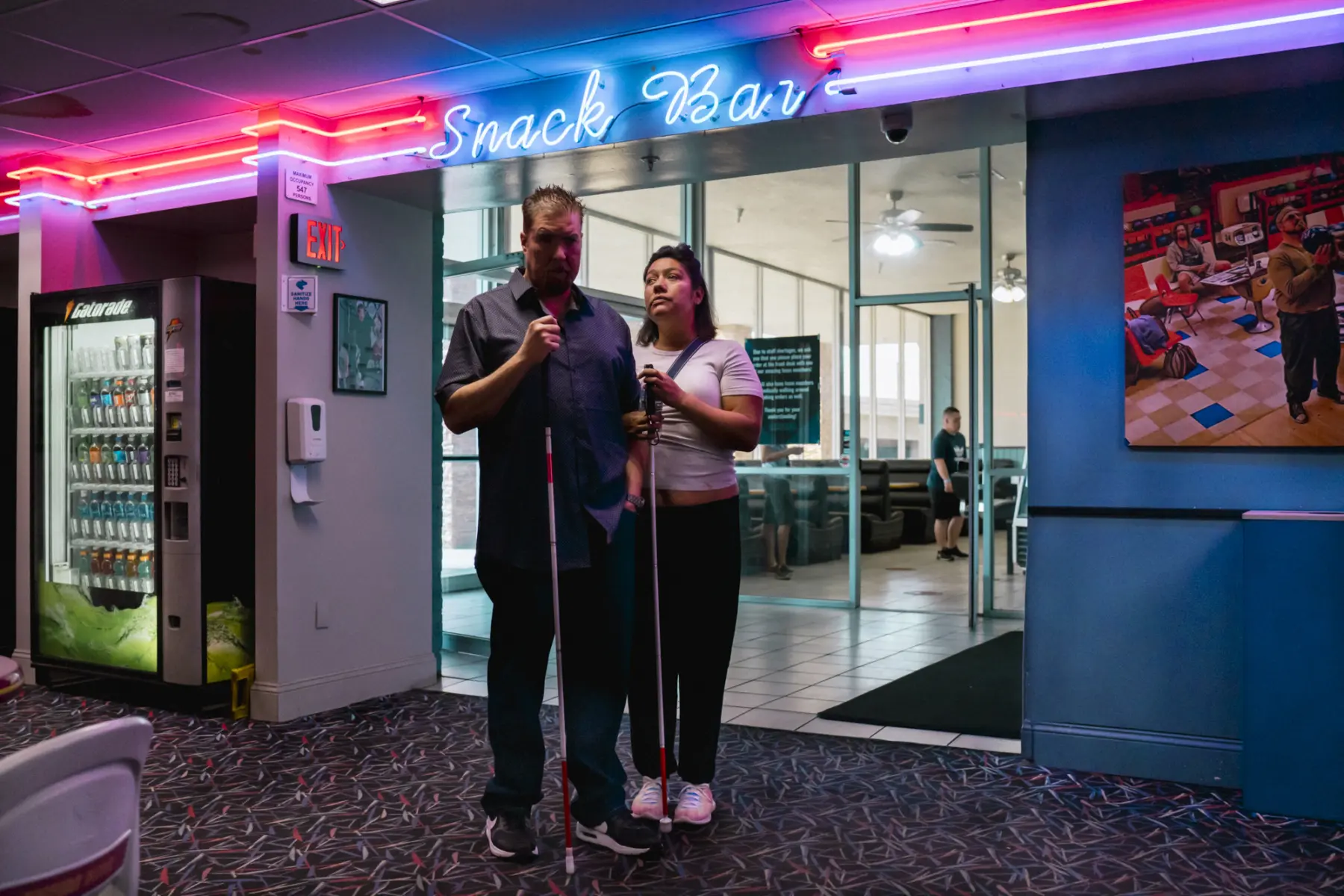 Melissa and Scott stand arm in arm holding their white canes beneath red, pink, and blue neon lights that illuminate their faces. The sign above them reads “snack bar.” A gatorade machine is on the left, and a poster of a man holding a bowling ball is on the right.