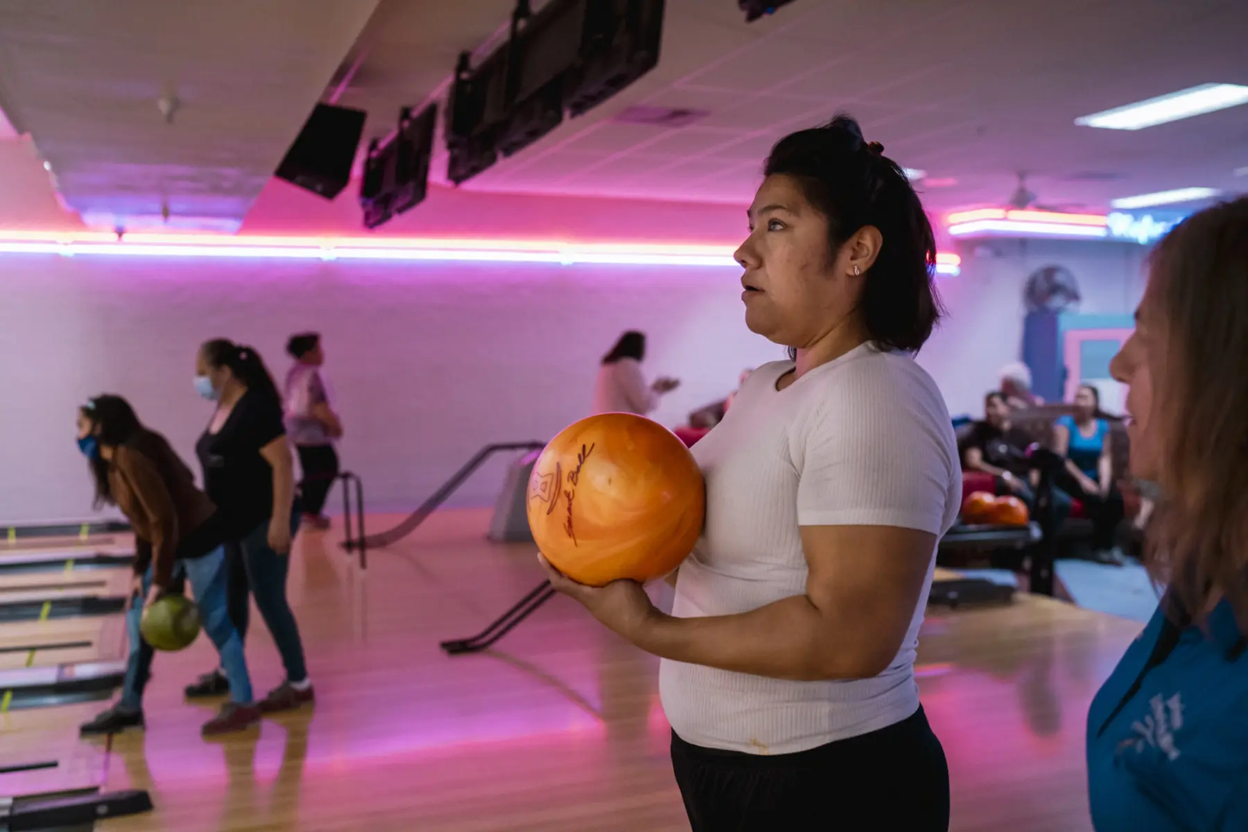 Melissa holds a bowling ball in front of a lane at a bowling alley and a woman who is out of focus is seen on the right looking over at Melissa. People who are out of focus bowl behind Melissa. There are pink neon lights in the background.