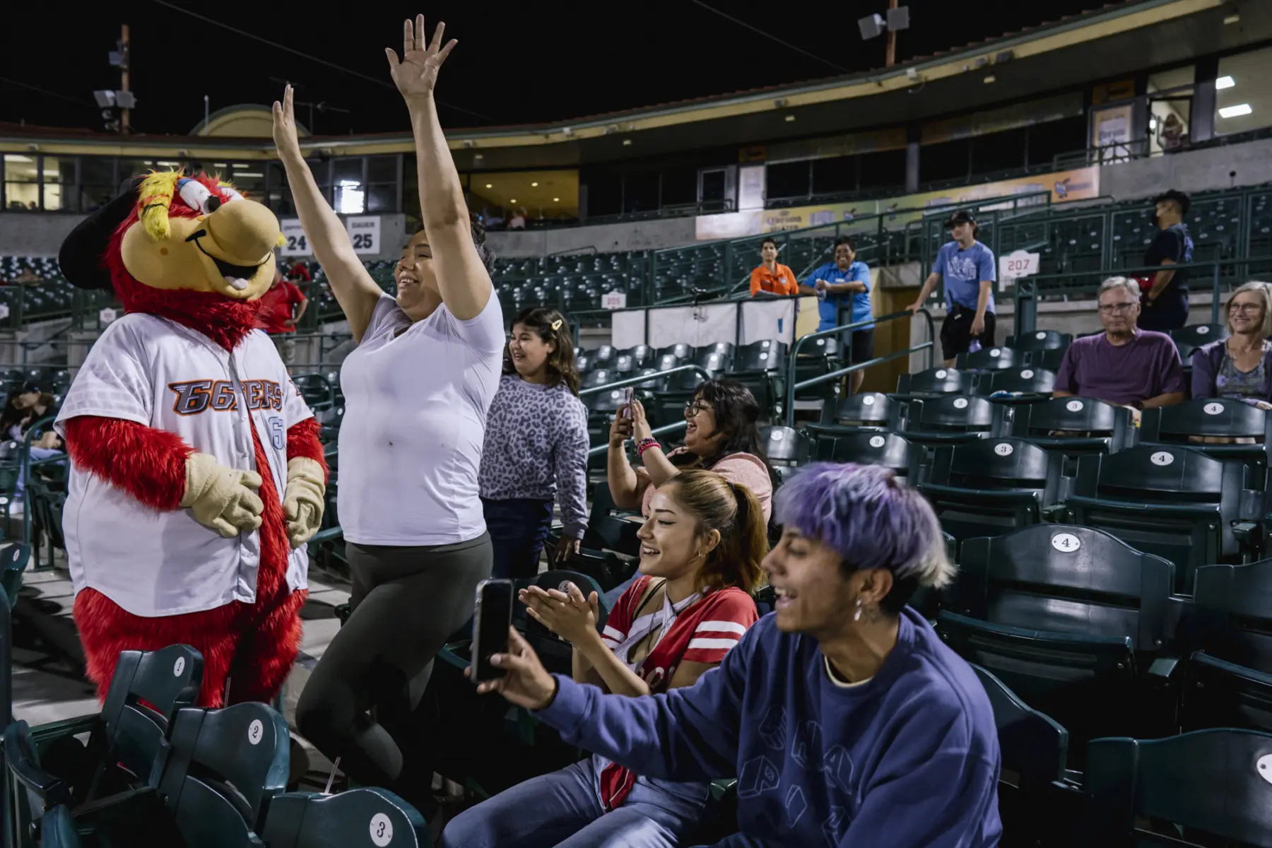In a sports arena, a smiling Melissa dances in front of her seat with arms in the air next to a bird mascot. People around her clap, take phone video, and look over with joyful facial expressions.
