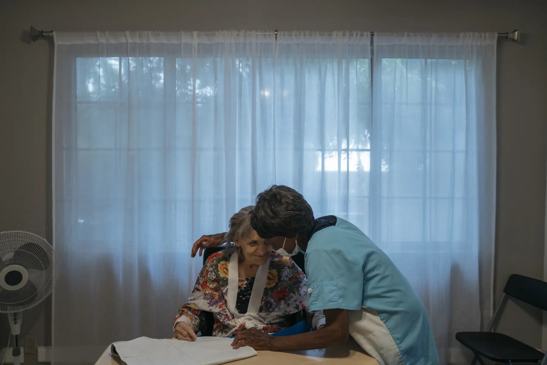 Ann, a white woman in her 70s with gray hair, sits smiling in her wheelchair at a dining room table with Mary, a Liberian woman in scrubs and a surgical mask. Behind them a blue hue emanates from closed sheer white drapes.