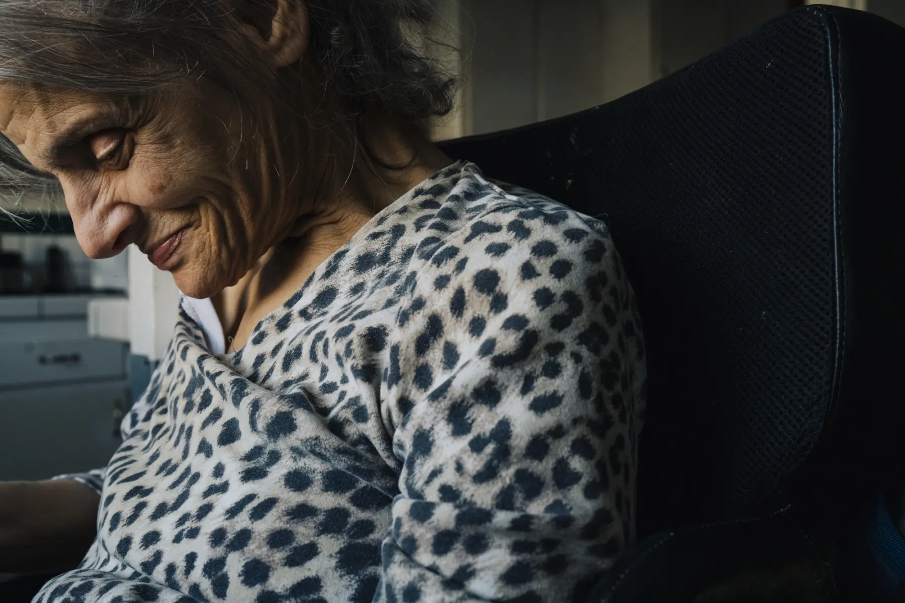 Ann sits smiling with head bowed and eyes looking down in a close up against the black background of her chair.