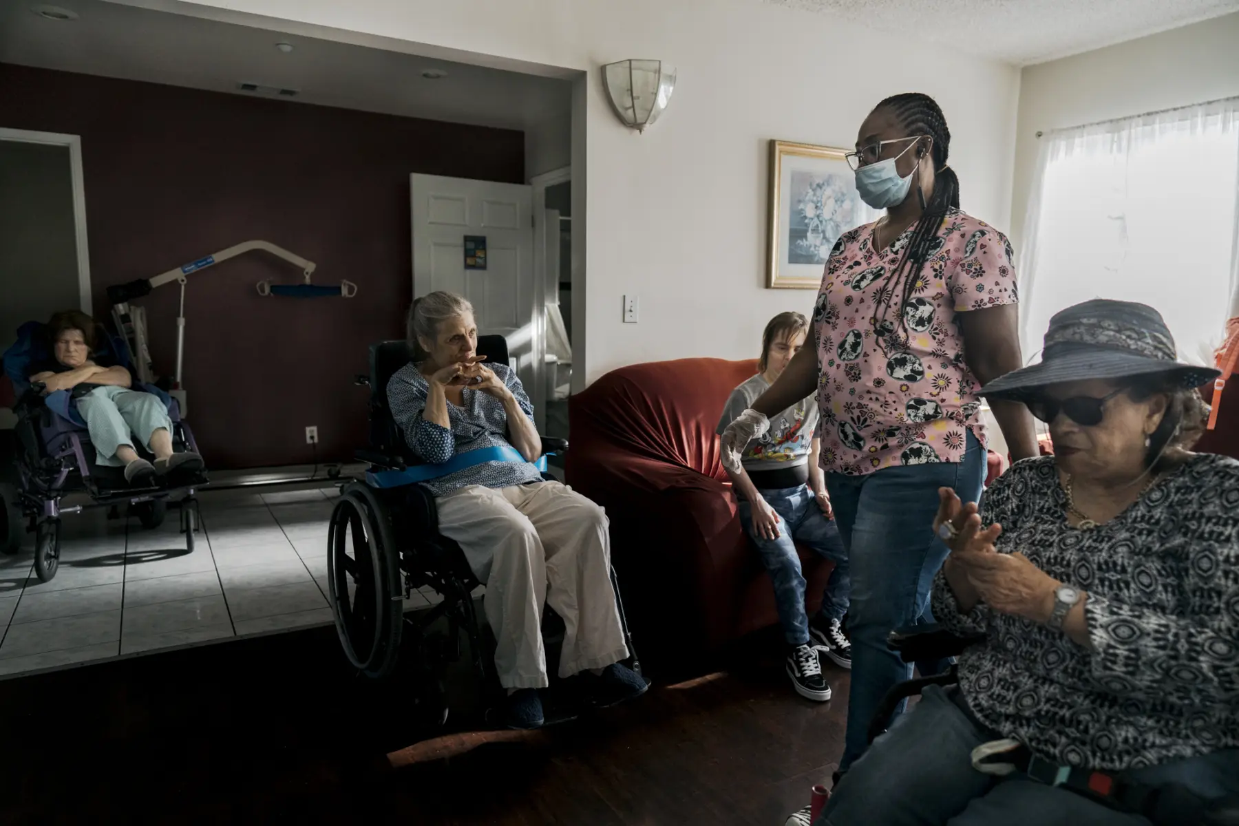 Ann sits in her wheelchair in a living room beside Juliet, a white woman in her 40s, who sits on a red velvet couch. Ann looks to Queen, a Black woman who wears scrubs and a surgical mask, walking by. In front of Queen, Norberta, a Hispanic woman in her 60s in a sunhat and large black sunglasses, sits in her wheelchair massaging her hands. In the far corner  Jill, a white woman in her 50s, reclines in her wheelchair with eyes closed and arms crossed.