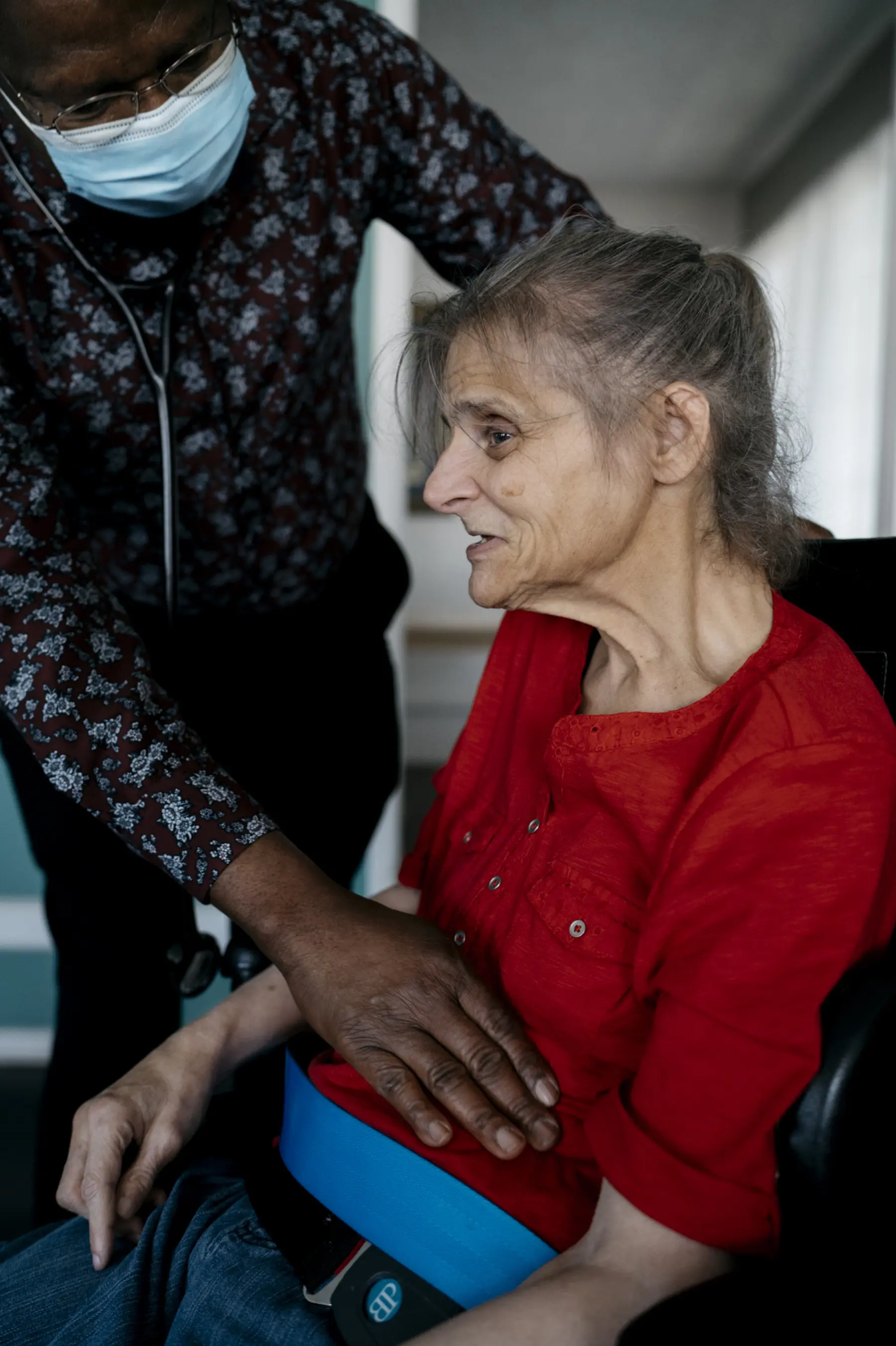 Nurse practitioner Dennis, an older Black male, leans over Ann and presses on her stomach with one hand. The nurse practitioner wears a surgical mask and has a stethoscope around his neck.