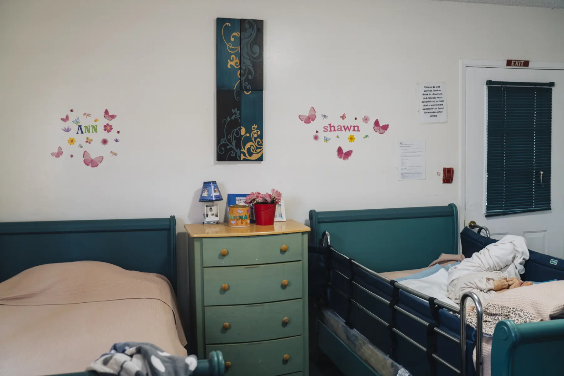 Two beds are on either side of a dresser that holds a small lamp, a Polaroid, a picture frame, and a flower pot. Ann’s bed is empty and her roommate Shawn lies in her own bed. On the wall above Ann’s bed, a sticker collage reads “Ann.” Above her roommate’s bed, stickers read “Shawn.”