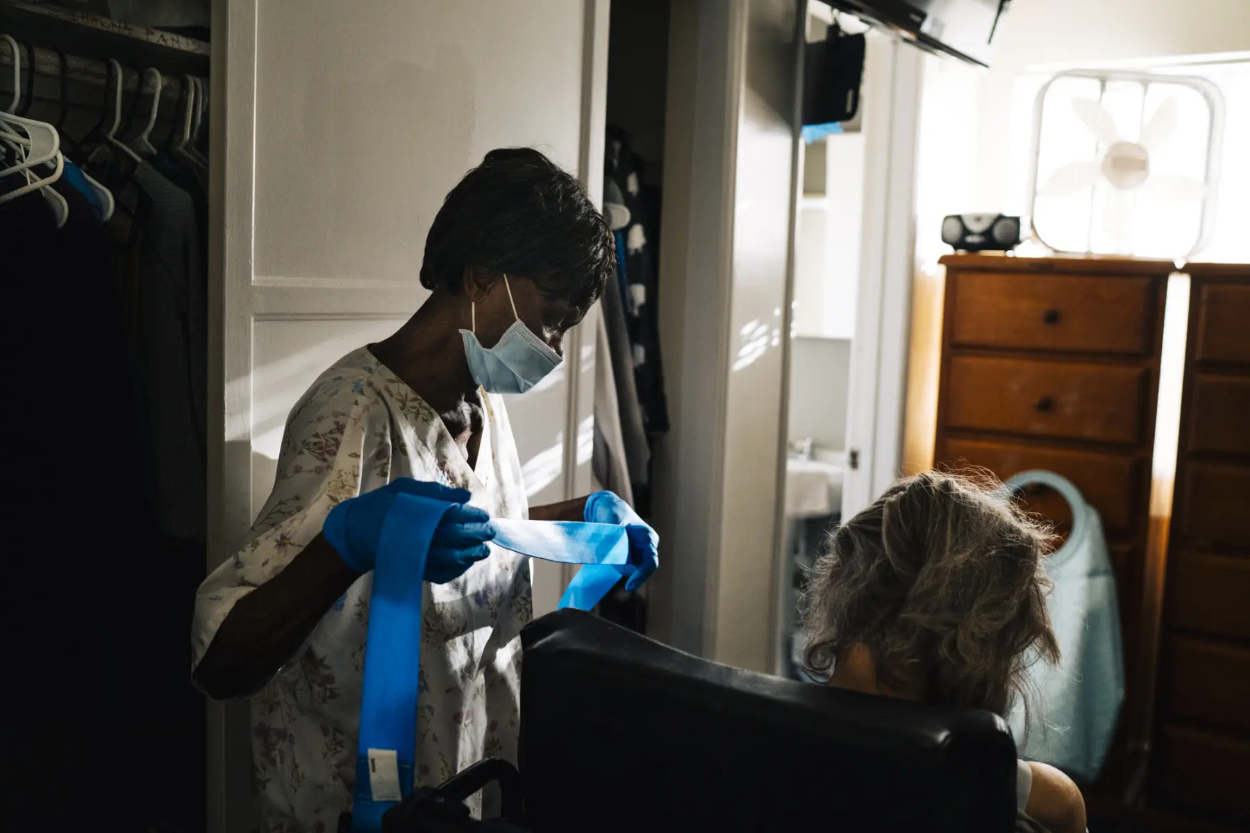 Mary, in scrubs and a surgical mask, stands beside Ann in front of Ann’s closet. Ann sits with only the back of her head seen. A soft light shines from the window in front of them, where a dresser with a small boombox and fan on top sits.