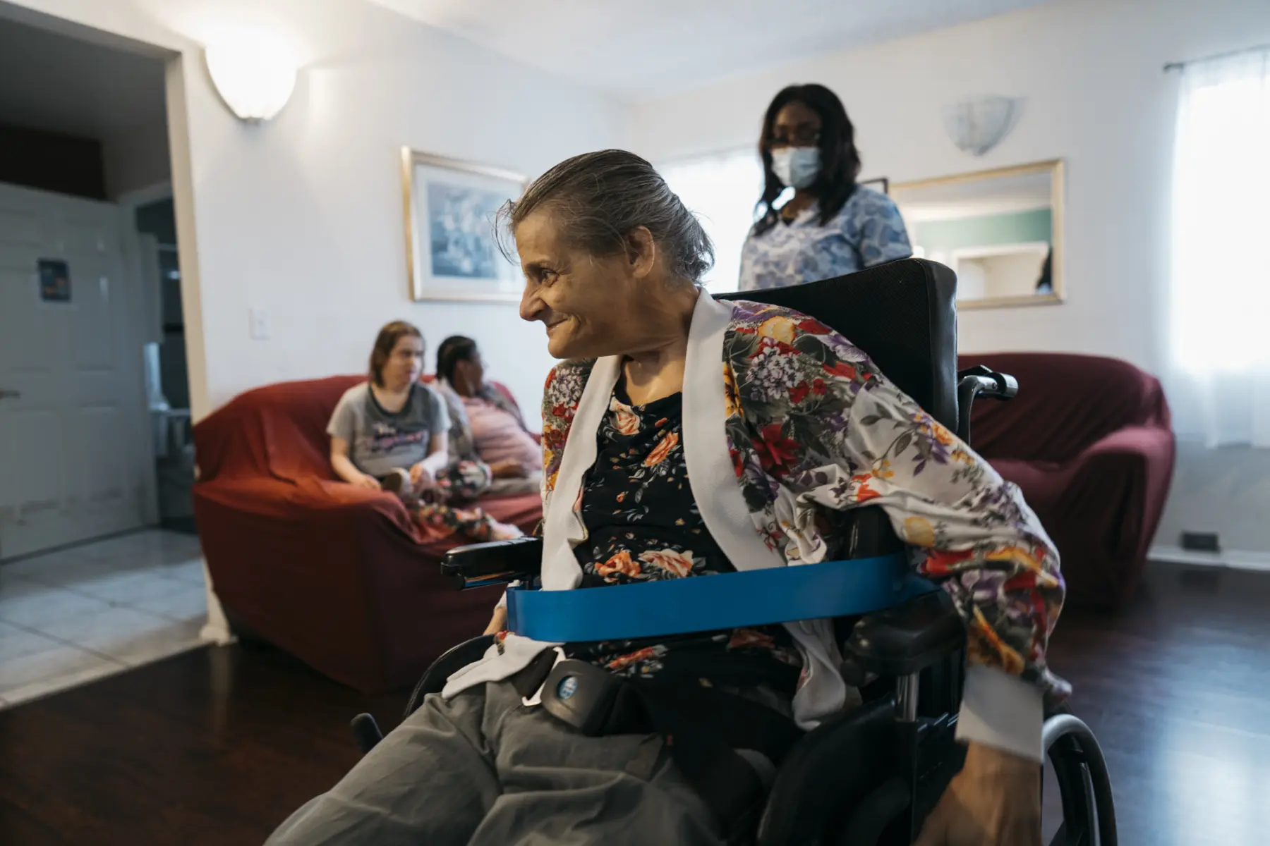 Ann looks to the side and smiles as she navigates the living room. Queen is out of focus behind Ann. On the couch behind Ann, also out of focus, sit Juliet and another housemate.