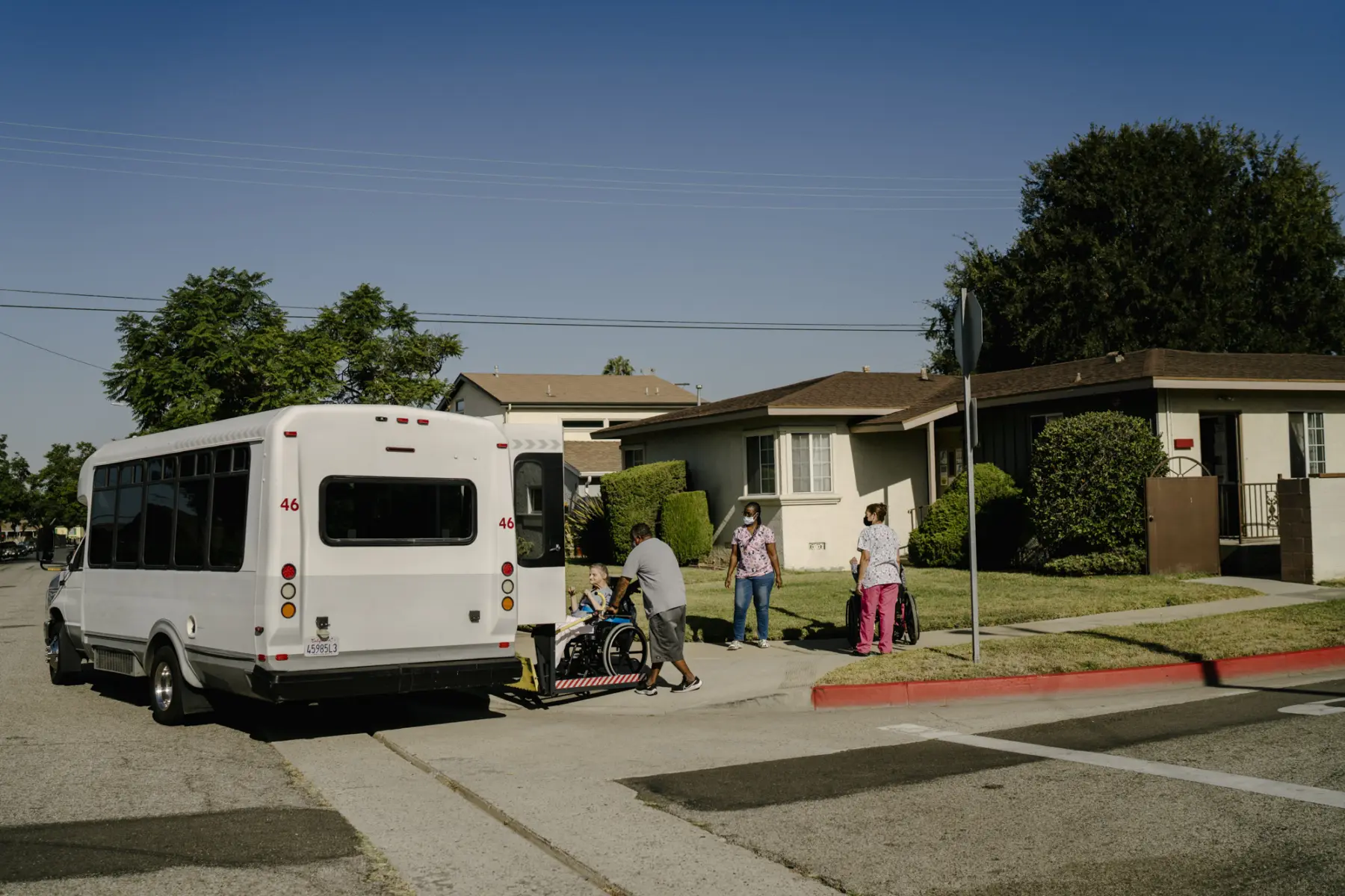 Steve, a Black male, pushes Ann in her wheelchair onto the ramp of a small transport bus parked in front of a single story home on a residential street. Two caregivers in scrubs wait with another resident, who sits in a wheelchair, under a blue sky.