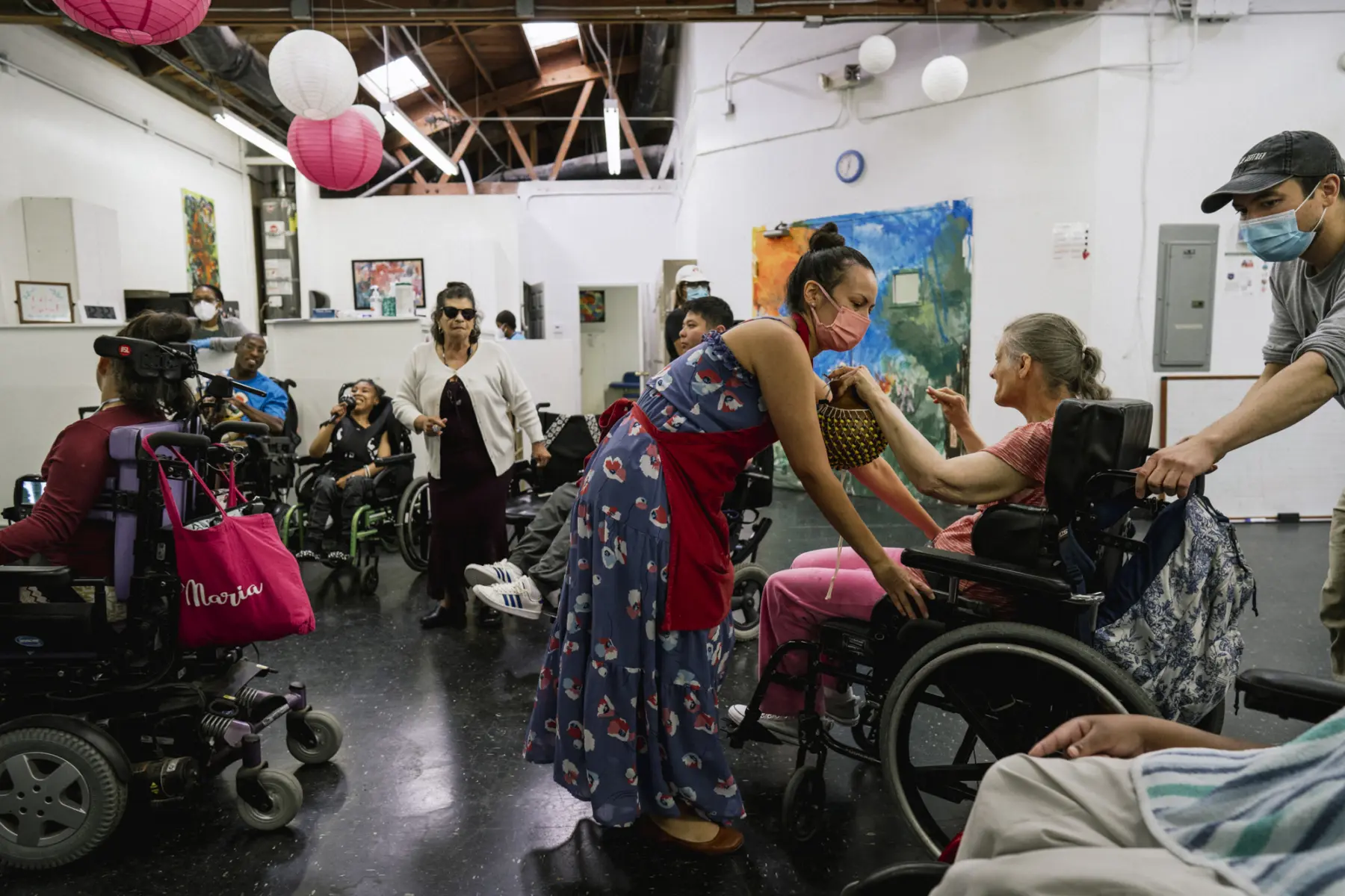 Stephanie, a Hispanic woman in her 30s, pulls Ann forward into group activity at a studio space. Jesse, a biracial Asian male in his 30s, pushes Ann from behind. Ann holds a beaded noise maker as she is maneuvered into a spot among a crowd of other artists who dance, sing, and hold maracas. Art fills the walls and lanterns hang from the ceiling.