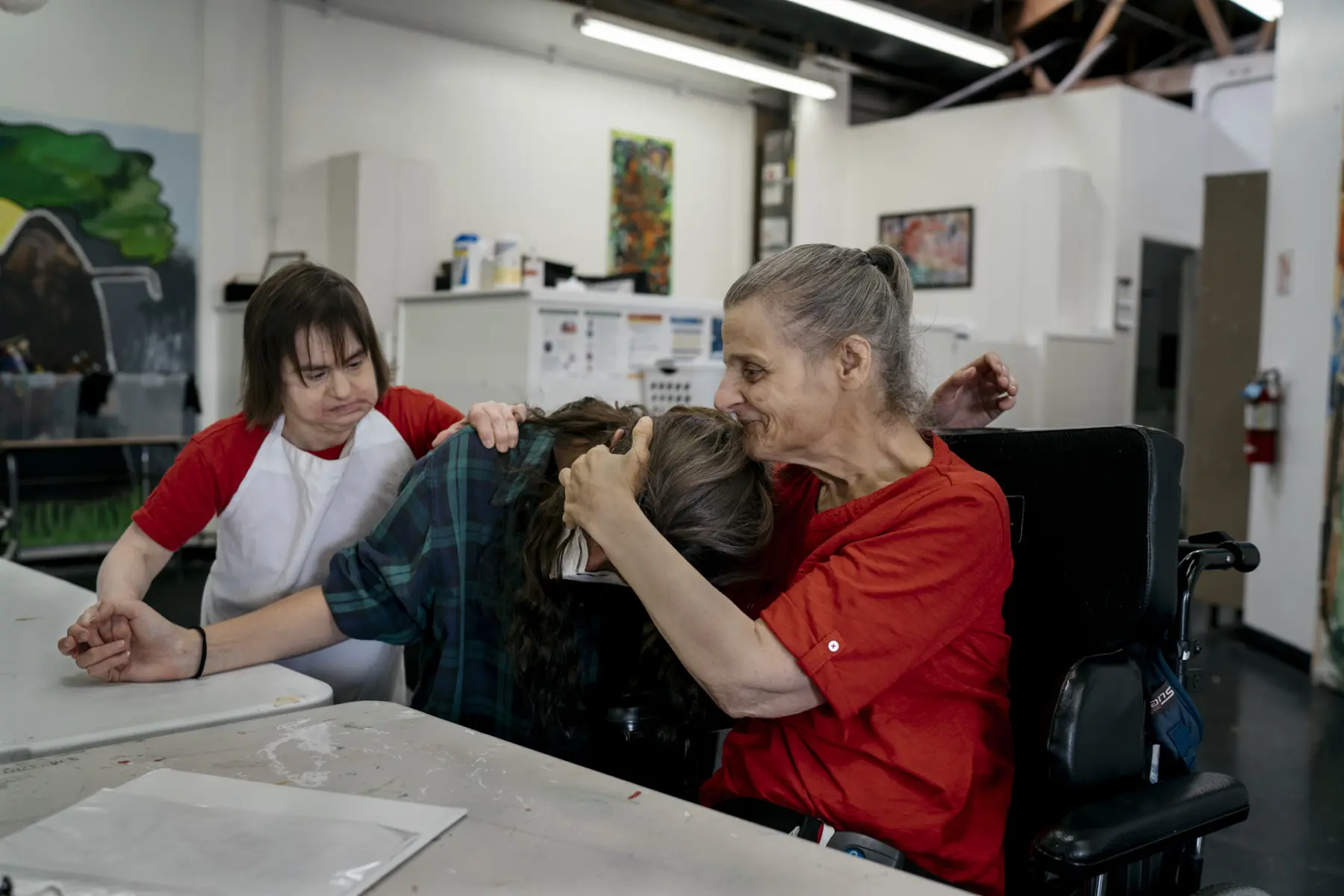 Ann smiles and embraces Dani, a white person with brown hair in her 20s, as they sit at a table in an art studio. Juliet, a white woman in her 40s, stands behind Dani with one hand on Dani’s shoulder and another holding Dani’s outstretched hand.