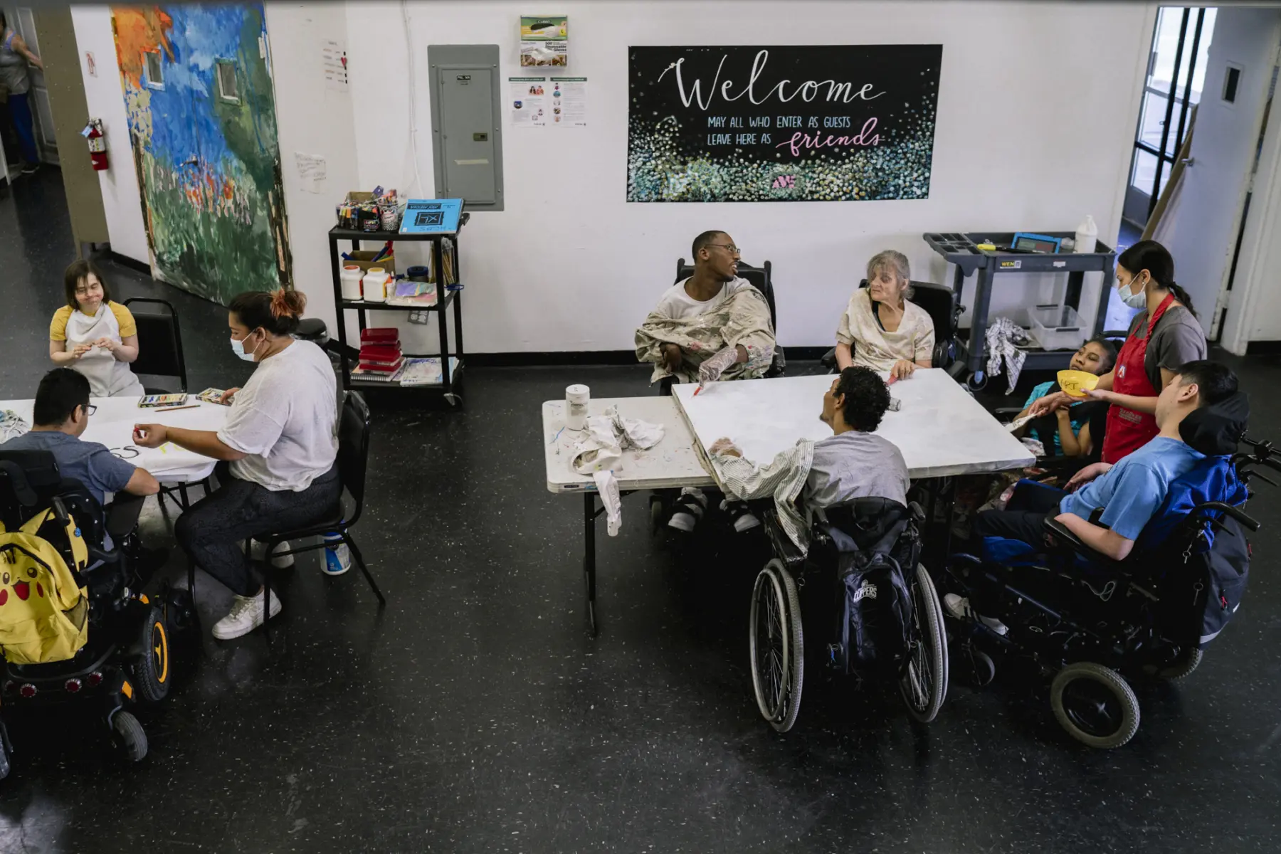 Ann as she sits at a table with a group of artists and an instructor as they paint together on a large white canvas. At another table at left, an instructor sits and draws with two participants. Behind them a poster reads “Welcome, May all who enter as guests leave here as friends.”