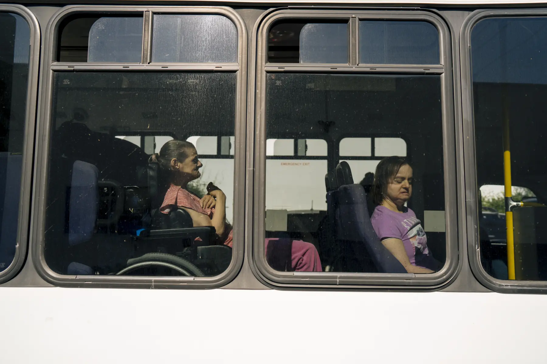 Ann is seen through the tinted windows of a transport bus behind friend Juliet. Sunlight illuminates both.