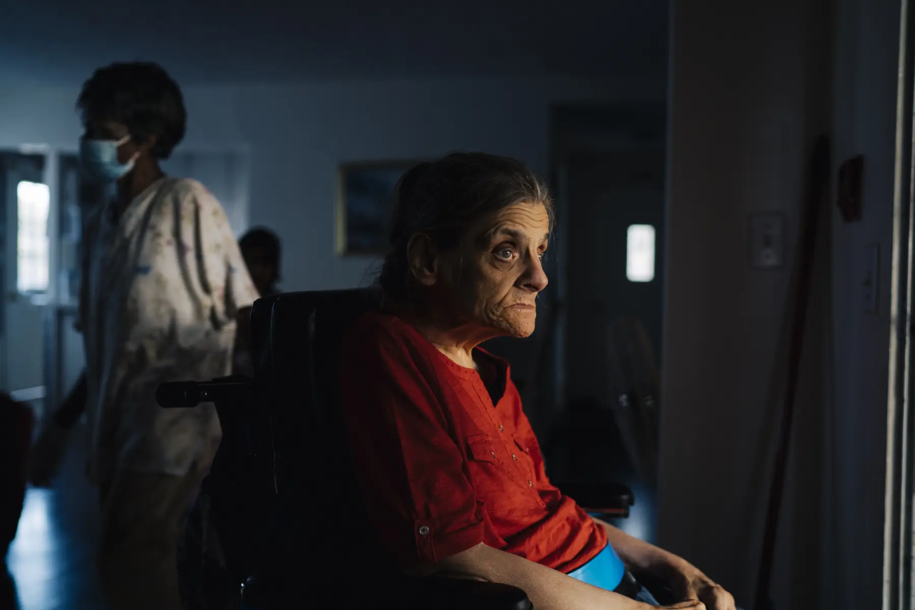 Ann sits in the living room illuminated by soft light. Mary is at left out of focus. A person is seen in shadow standing behind Mary.