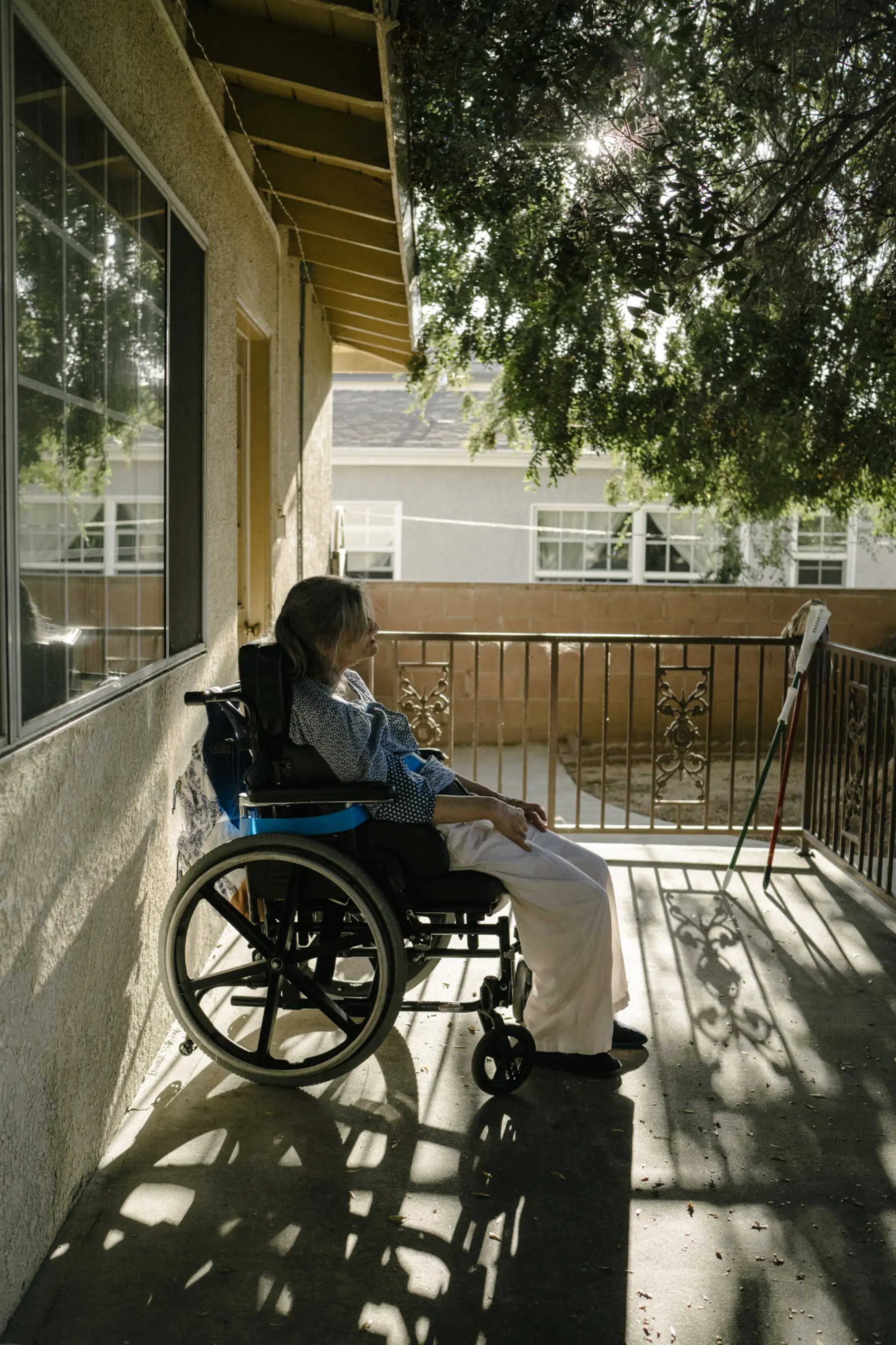 Ann sits on the back patio as light shines through the leaves on a tree. The shadows of a fence and her wheelchair are projected around Ann on the concrete. Another home is seen in the background.