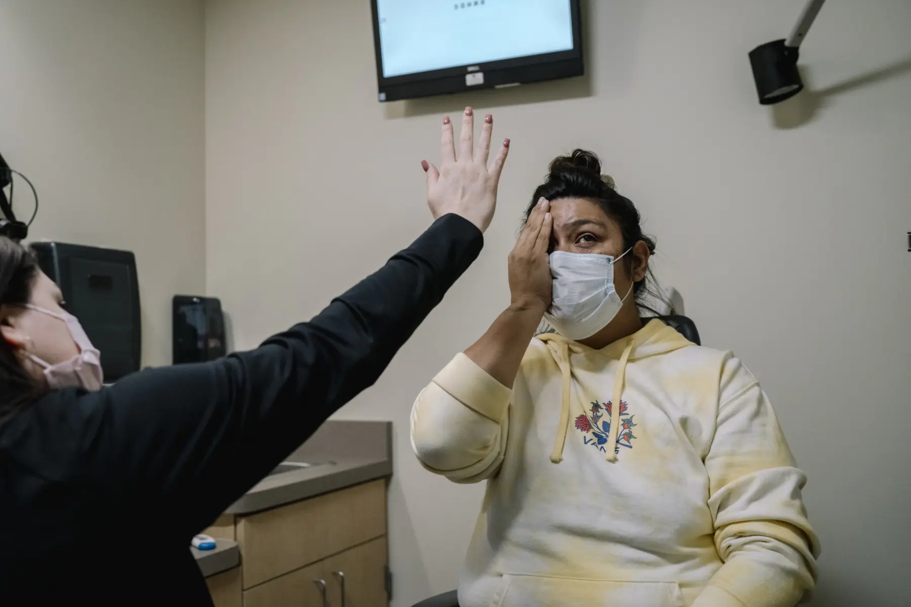 Melissa covers her left eye with her hand as she sits in an eye clinic. A nurse holds up her hand showing five fingers close to Melissa’s face. The nurse is only seen from behind. Both Melissa and the nurse wear surgical masks.