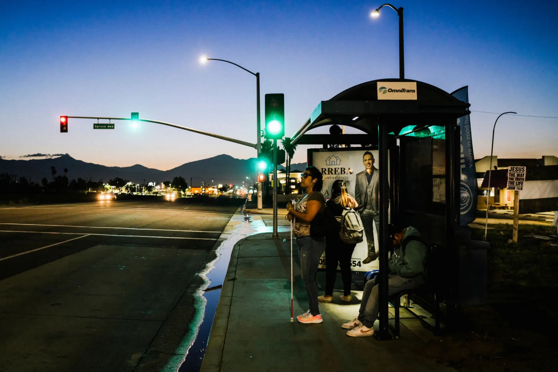 Melissa stands, holding her white cane, at a bus stop shelter next to an empty street and traffic light. Her teenage children, Angie and Jacob, stand and sit beside her wearing backpacks. The sun rises over the outline of a mountain range behind them.