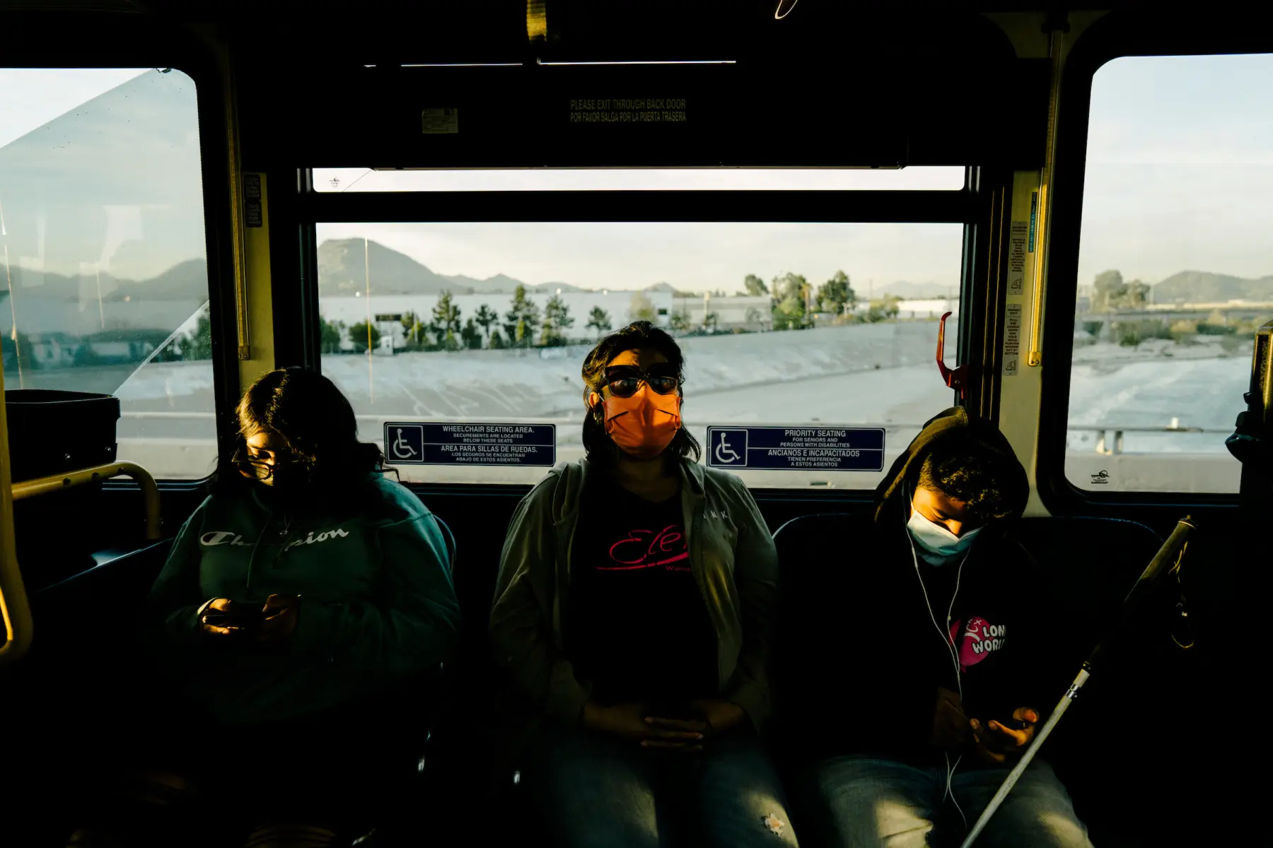 Melissa wears sunglasses and listens to music on earbuds between Angie and Jacob, who look at their phones.The landscape and mountain range are seen through a window behind them. Light hits their faces.