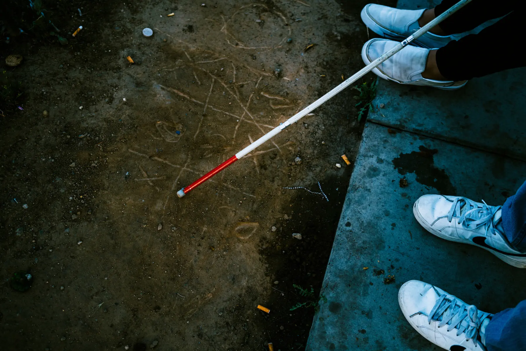Melissa’s white cane rests on a game of tic-tac-toe etched into dirt next to a concrete sidewalk, where Angie’s and Jacob’s sneakers are seen side-by-side.