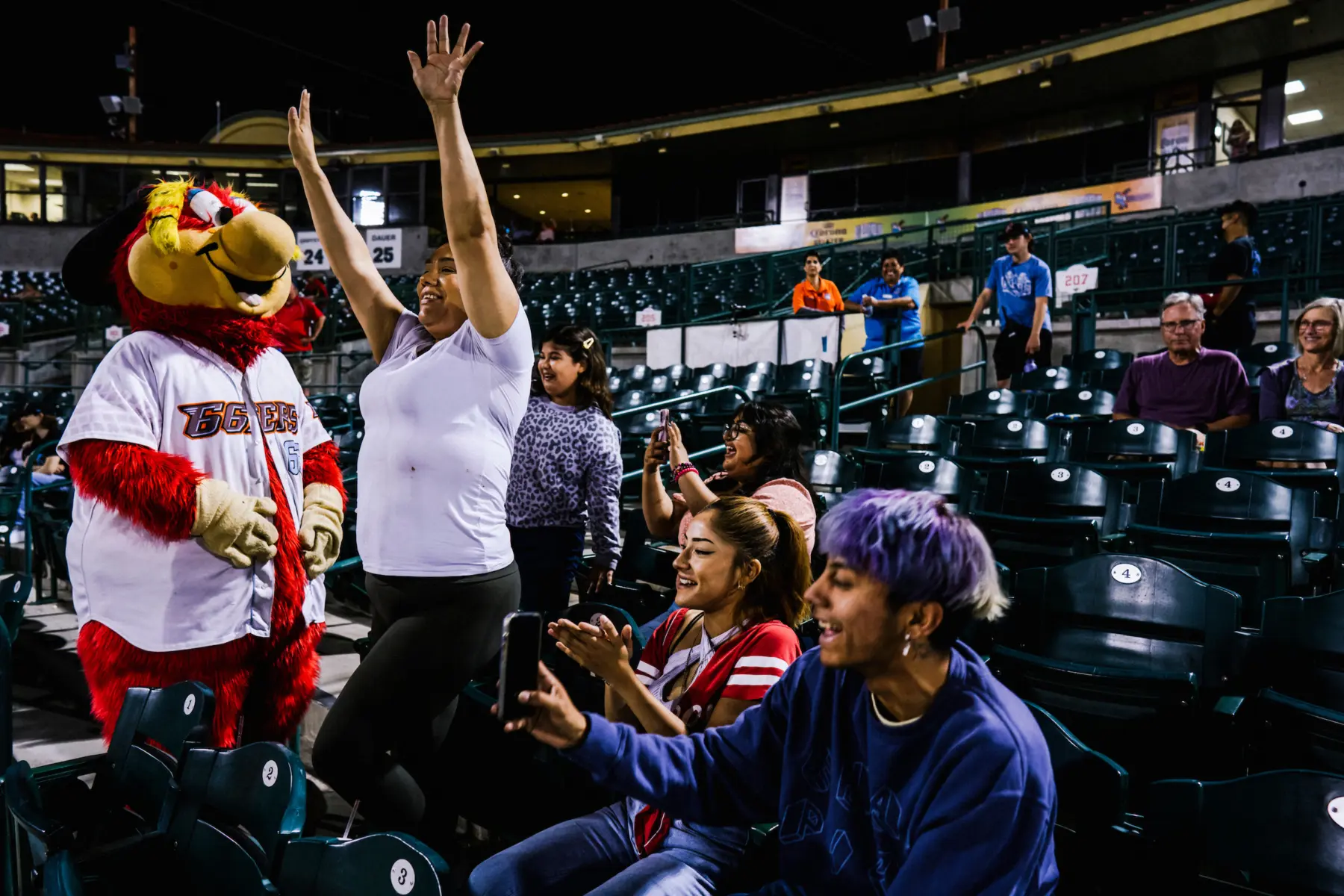 In a sports arena, a smiling Melissa dances in front of her seat with arms in the air next to a bird mascot. People around her clap, take phone video, and look over with joyful facial expressions.