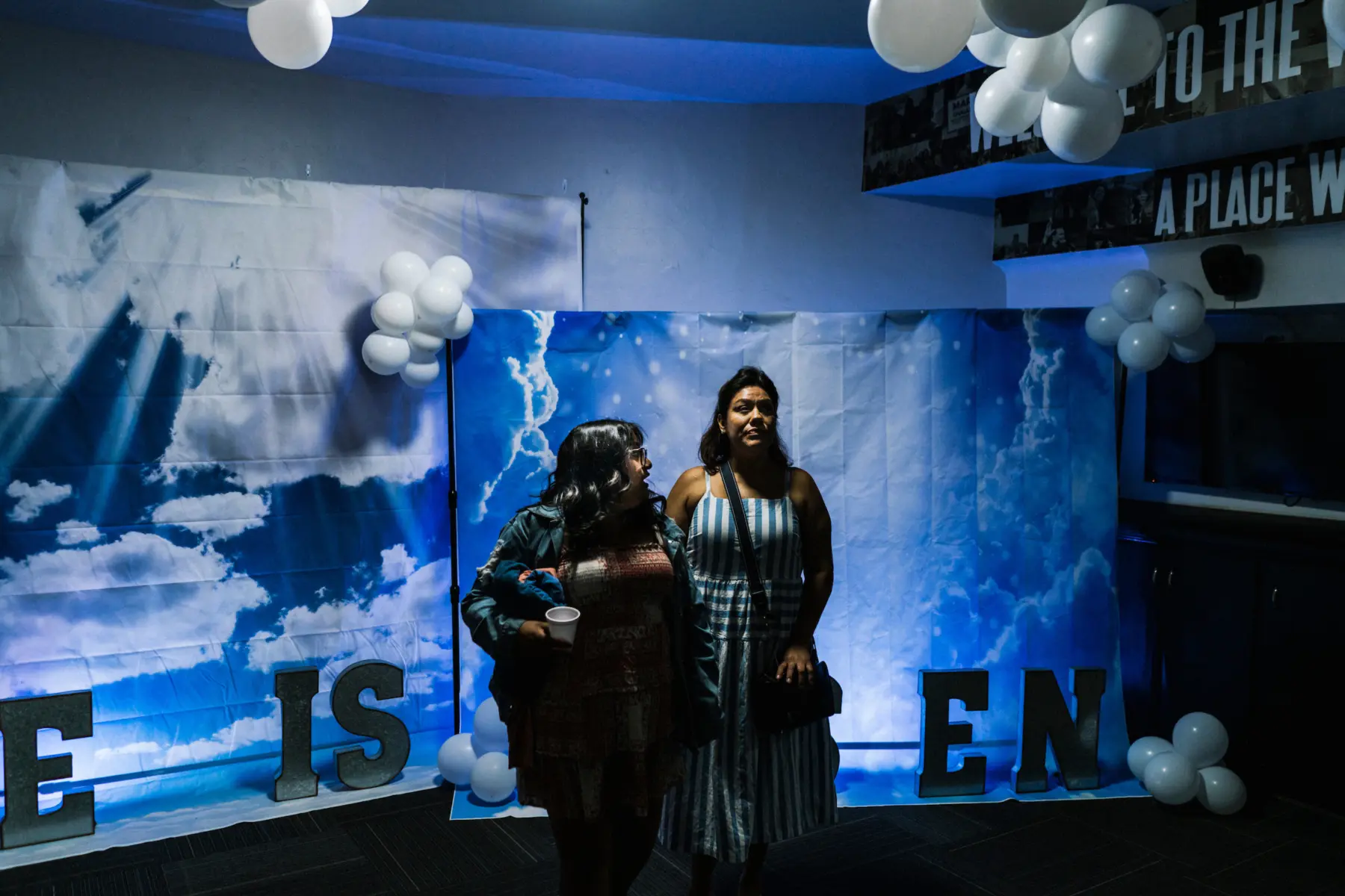 Angie looks to her mother, Melissa, as they walk arm in arm in front of backdrops of blue skies and large clouds. A portion of large letters below the backdrops read “He is risen.” White balloons hang from the ceiling and from the backdrops.