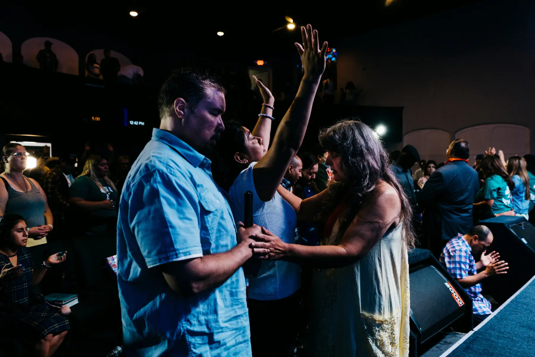 Melissa holds her hands in the air and closes her eyes as she receives a prayer at a church altar. Scott stands behind Melissa with his eyes closed. Another woman stands in front of Melissa with her eyes closed and one hand on Melissa’s shoulder and one hand on Scott’s hand. Behind them, a crowd of people pray on the ground floor and balcony.