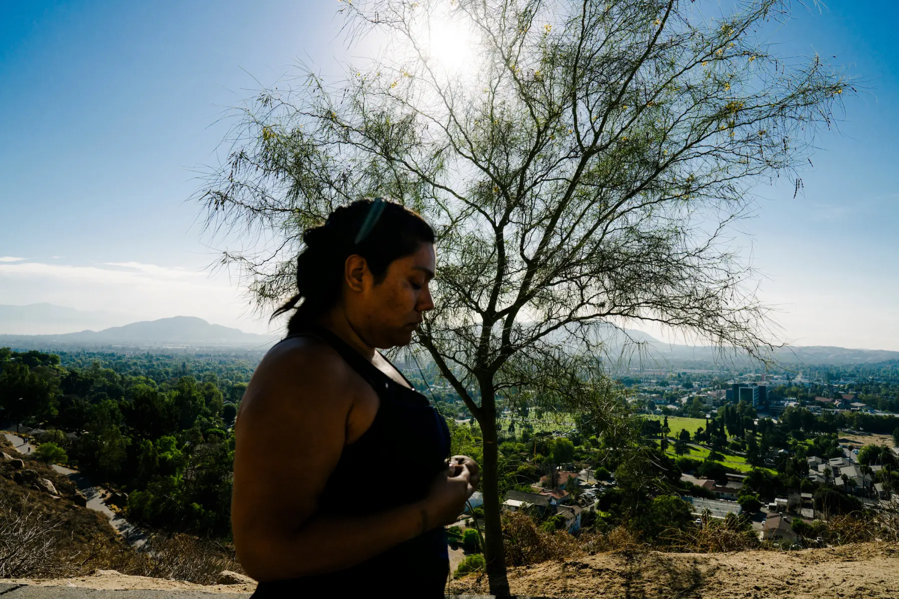 Melissa is seen from the side as she stands at a high elevation point overlooking a city landscape. Behind her is a tree. Melissa is out of focus, while the tree and the city landscape are in focus.