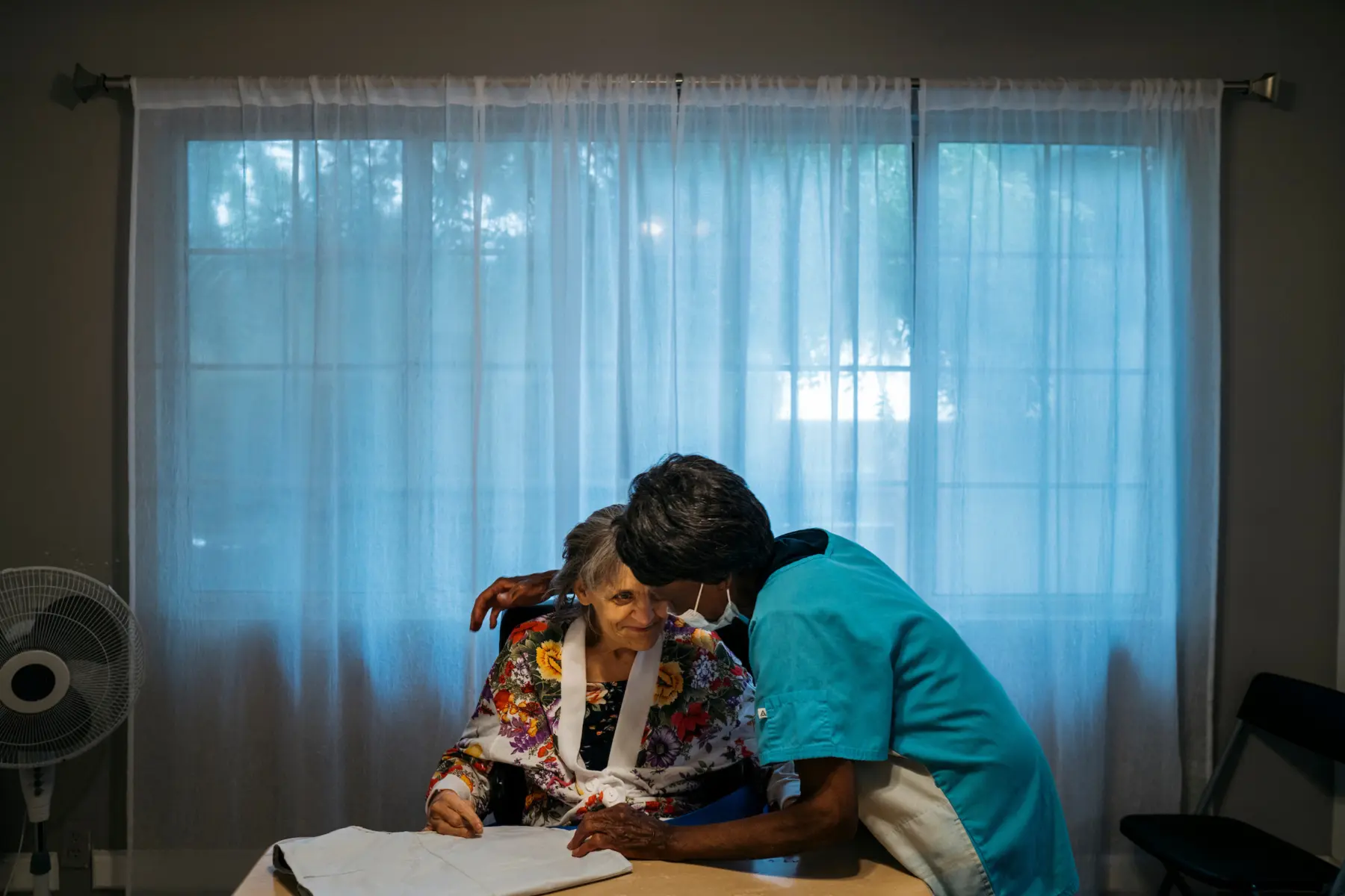 Ann, a white woman in her 70s with gray hair, sits smiling in her wheelchair at a dining room table with Mary, a Liberian woman in scrubs and a surgical mask. Behind them a blue hue emanates from closed sheer white drapes.