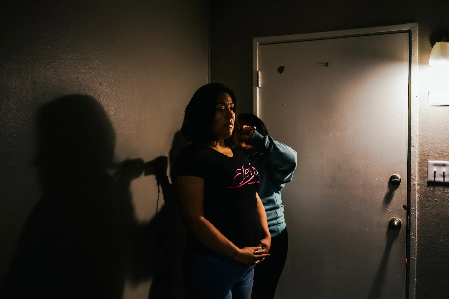 Melissa stands by a door while Angie stands behind her blow drying her hair. Shadows of Melissa’s figure and Angie’s raised arm are seen on the wall beside them.