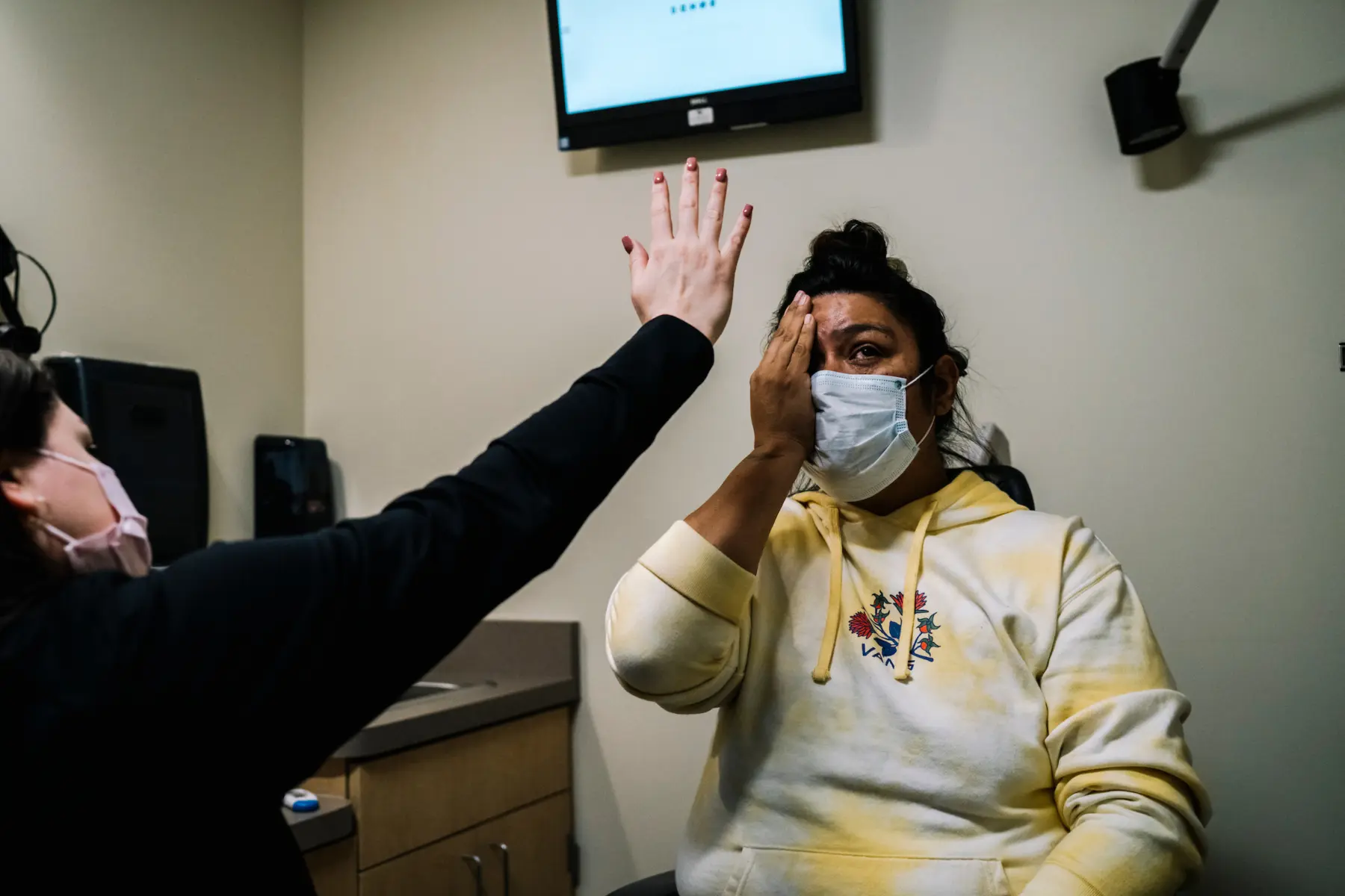 Melissa covers her left eye with her hand as she sits in an eye clinic. A nurse holds up her hand showing five fingers close to Melissa’s face. The nurse is only seen from behind. Both Melissa and the nurse wear surgical masks.