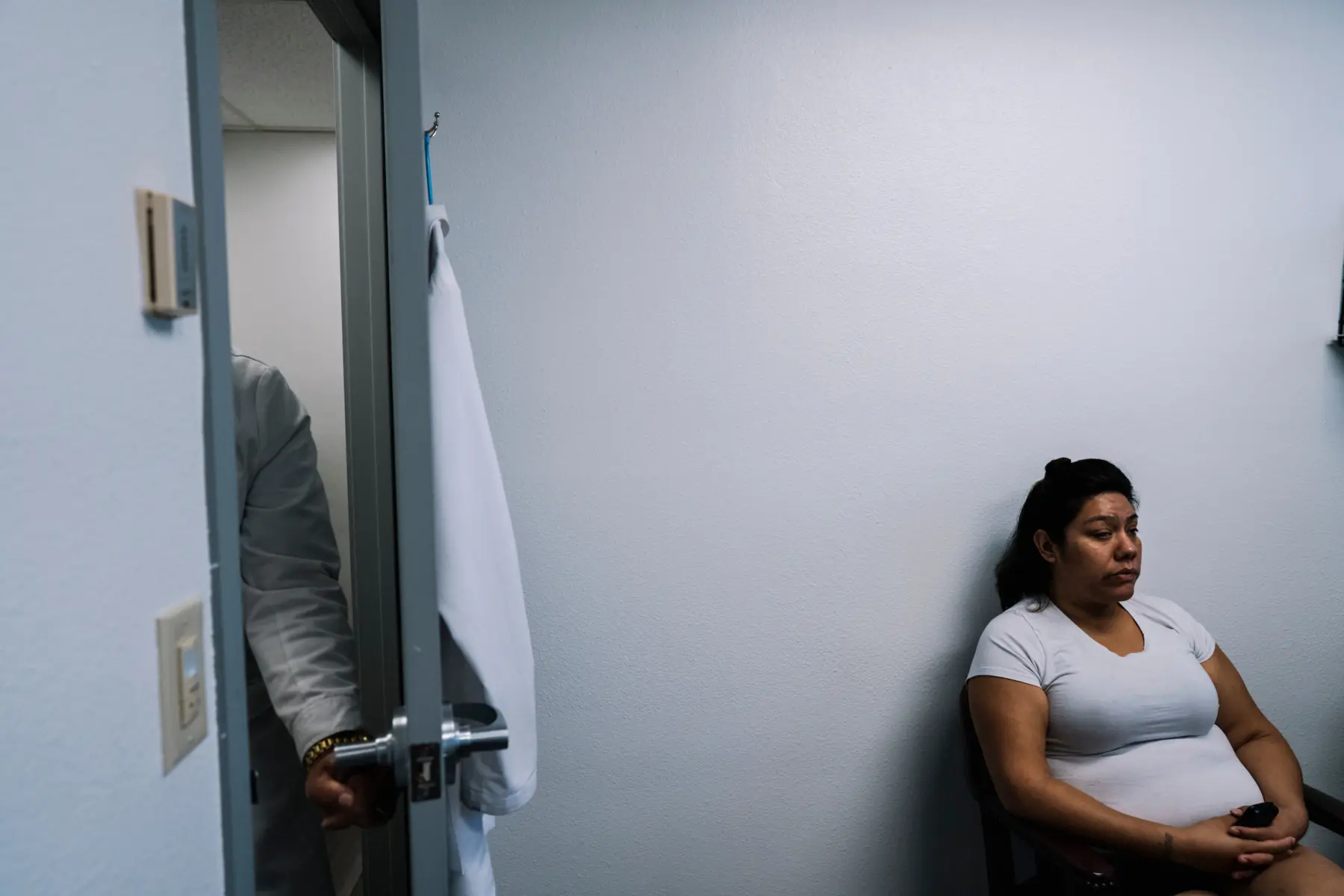 Melissa sits patiently in a doctor’s office against a stark white wall. A door opens to the left revealing only a doctor’s arm pushing the door open toward her.